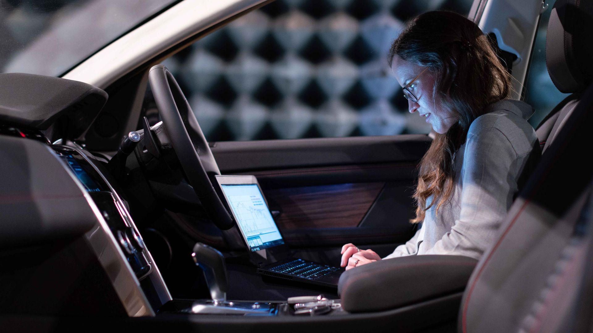 Woman working on a laptop inside a car, testing sound engineering in an isolated chamber.