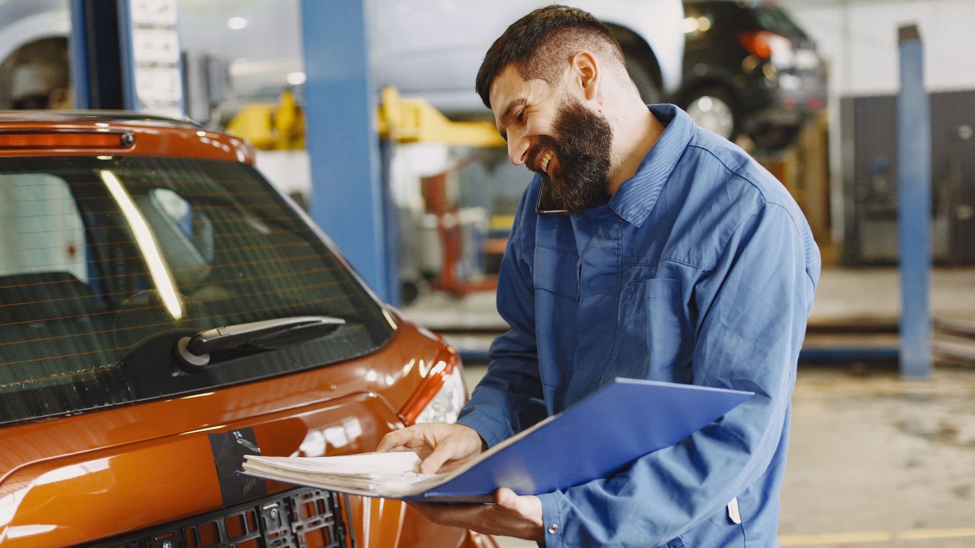 Smiling auto mechanic inspects a car in a service garage, wearing coveralls and holding documents.