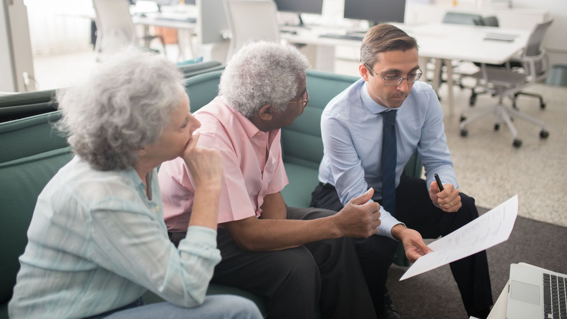 Business professional consults elderly clients in an office setting. Collaborative discussion, paperwork visible.