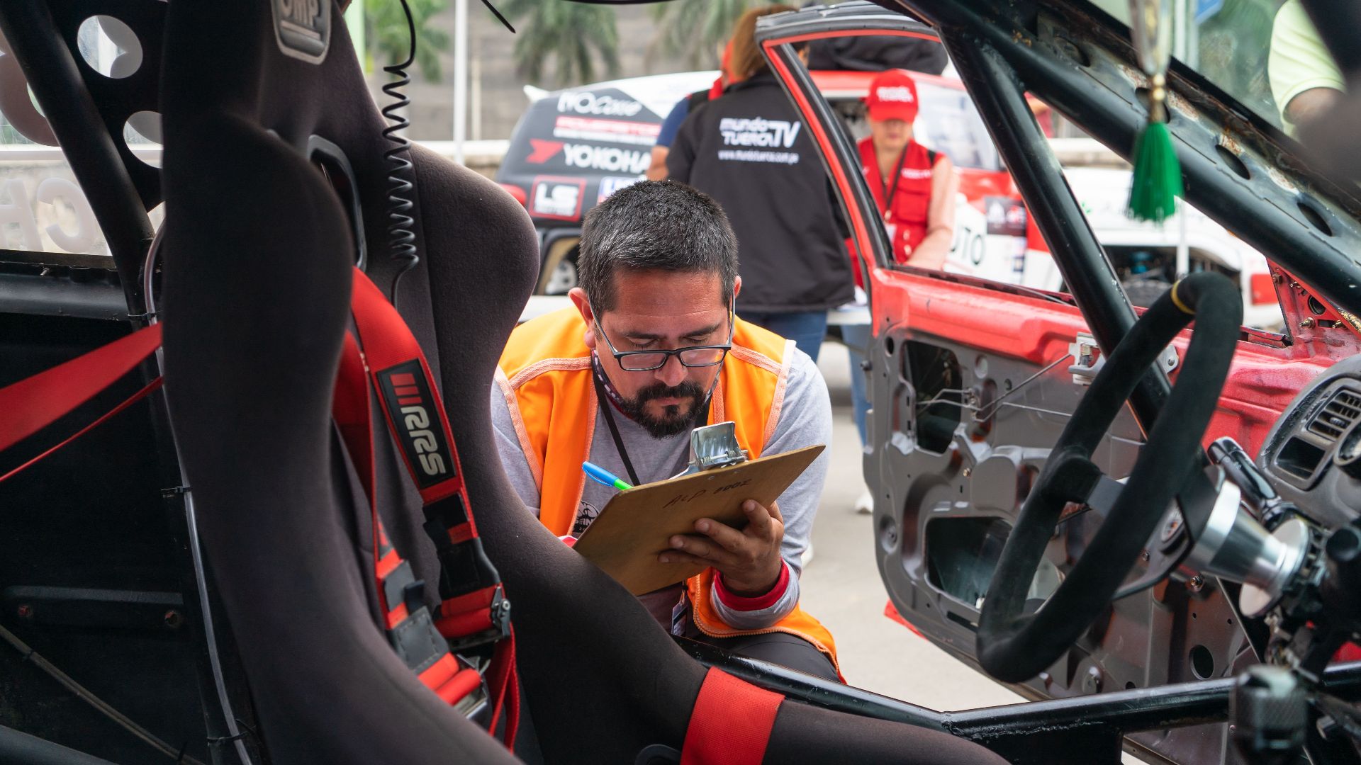 A man inspects a race car interior at a motorsport event, noting findings on a clipboard.