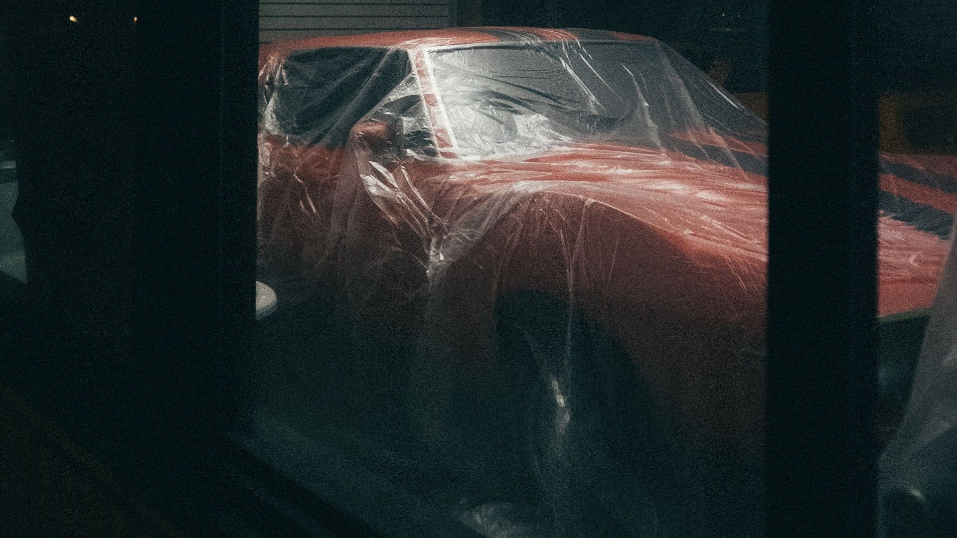 A dimly lit indoor scene showcasing a classic red car covered with protective plastic.
