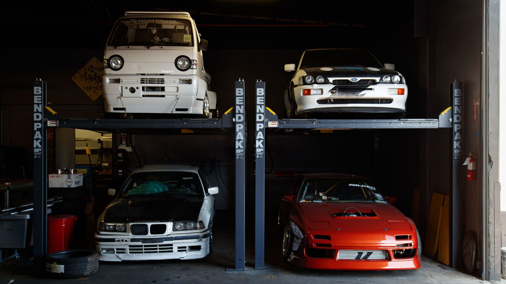 A collection of modified sports cars on a mechanical garage lift in Compton, CA.