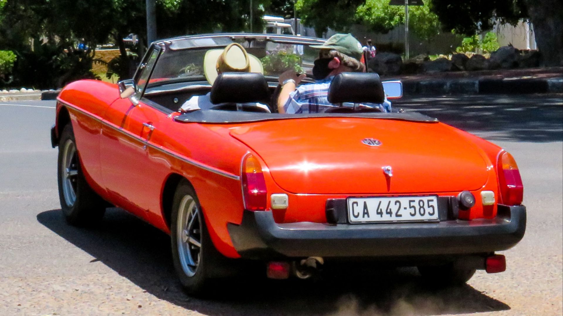 A vibrant red classic convertible driving outdoors on a sunny day with two adults enjoying the ride.