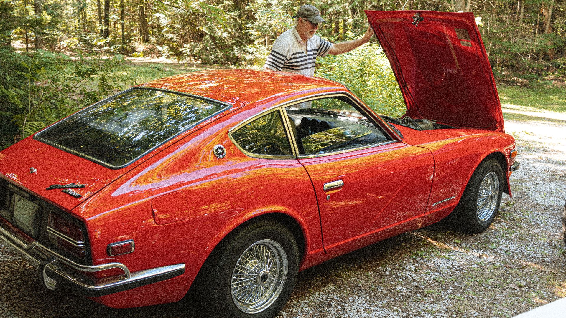 A senior man inspects the engine of a classic red Datsun 240Z parked outdoors.