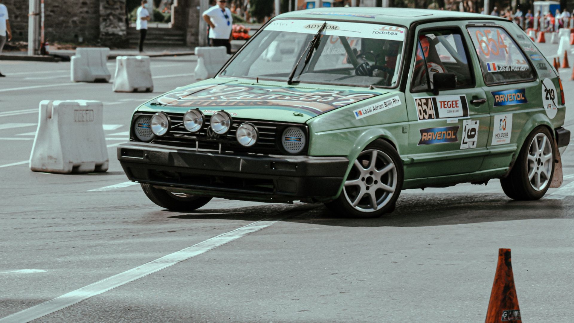 Green retro automobile with stickers riding on road with marking lines near on autodrome with cones and barriers in street