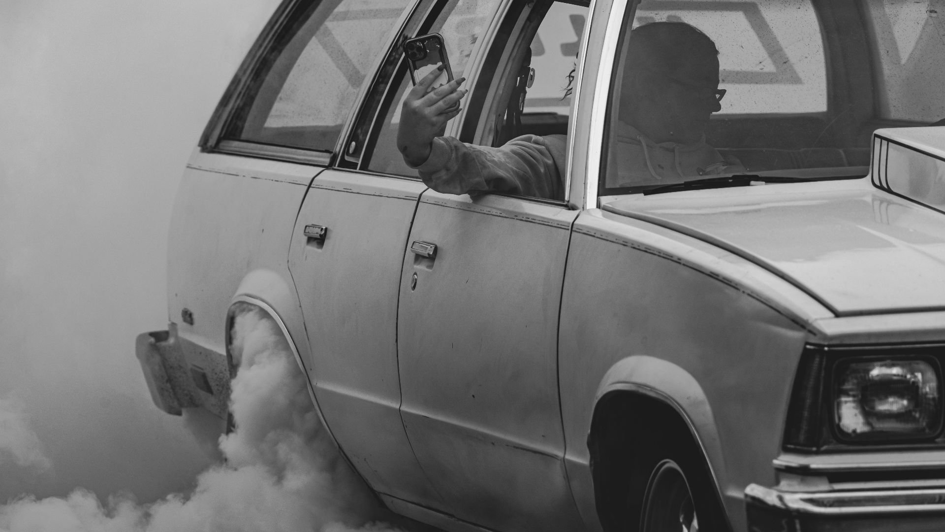 Black and white image of a vintage car performing a smoky burnout with a man taking a selfie.