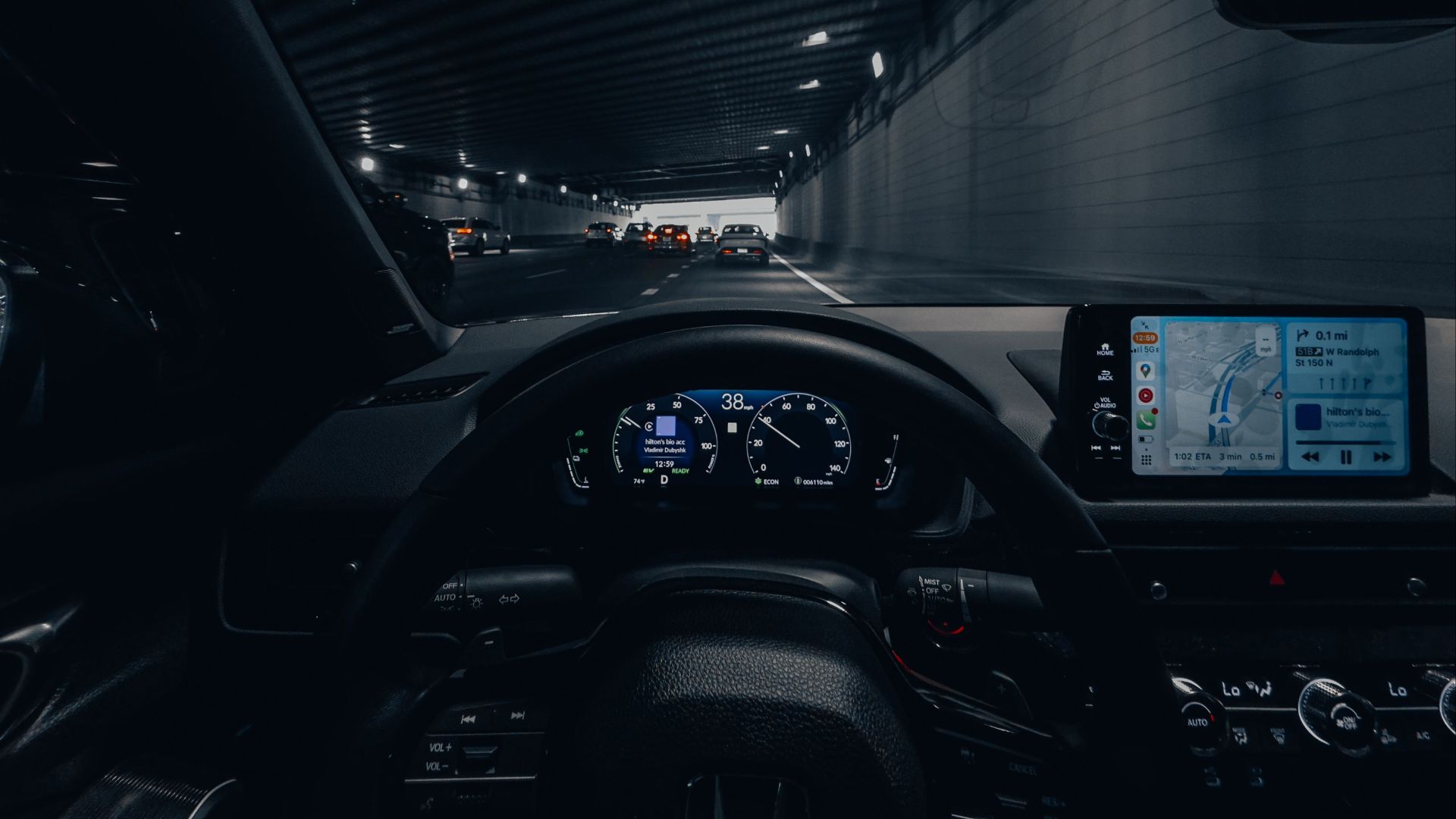 View from car dashboard driving through an illuminated city tunnel at night.