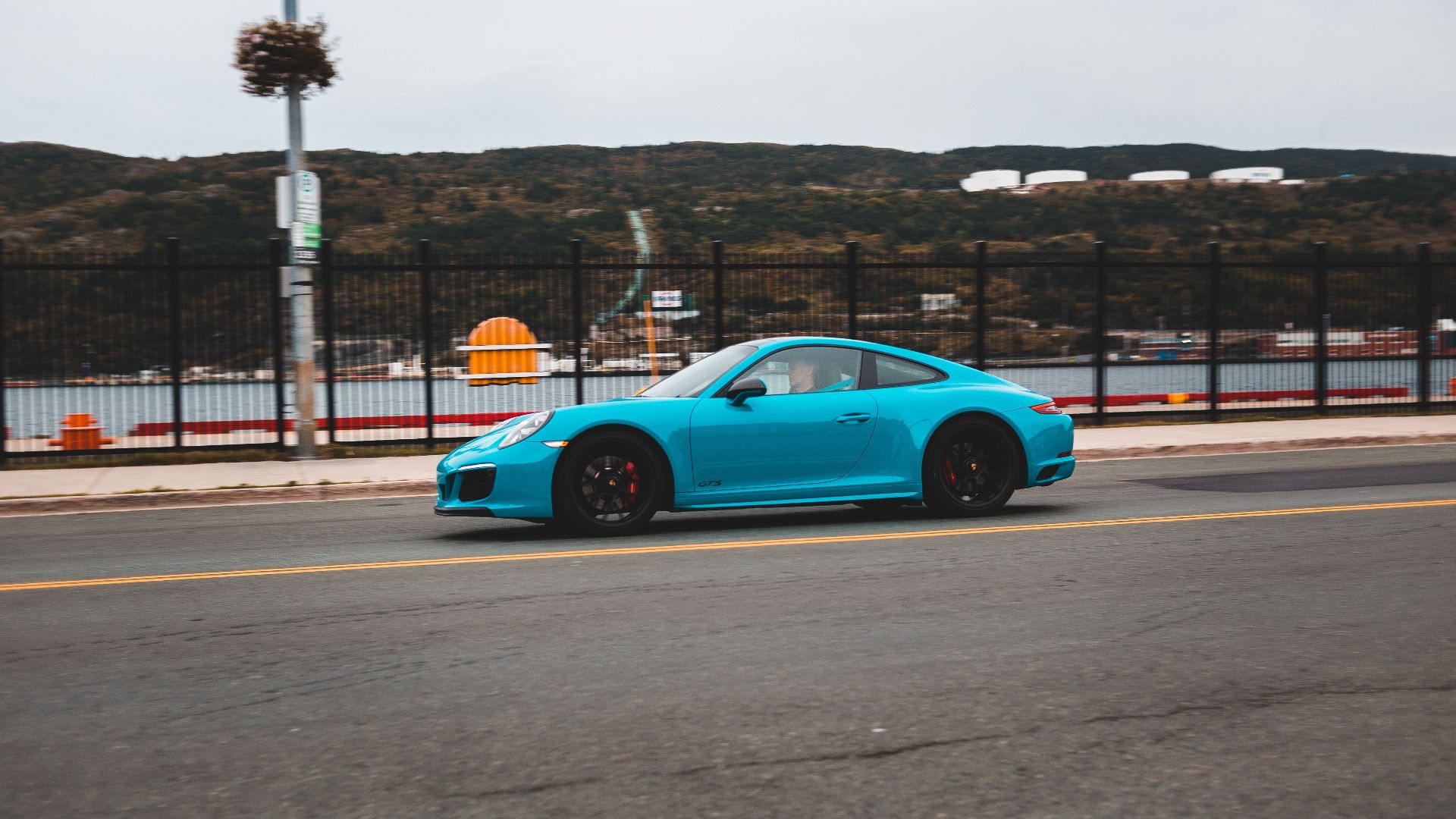 Side view of contemporary blue car driving on asphalt roadway near metal fence along embankment with sea with mountains in distance