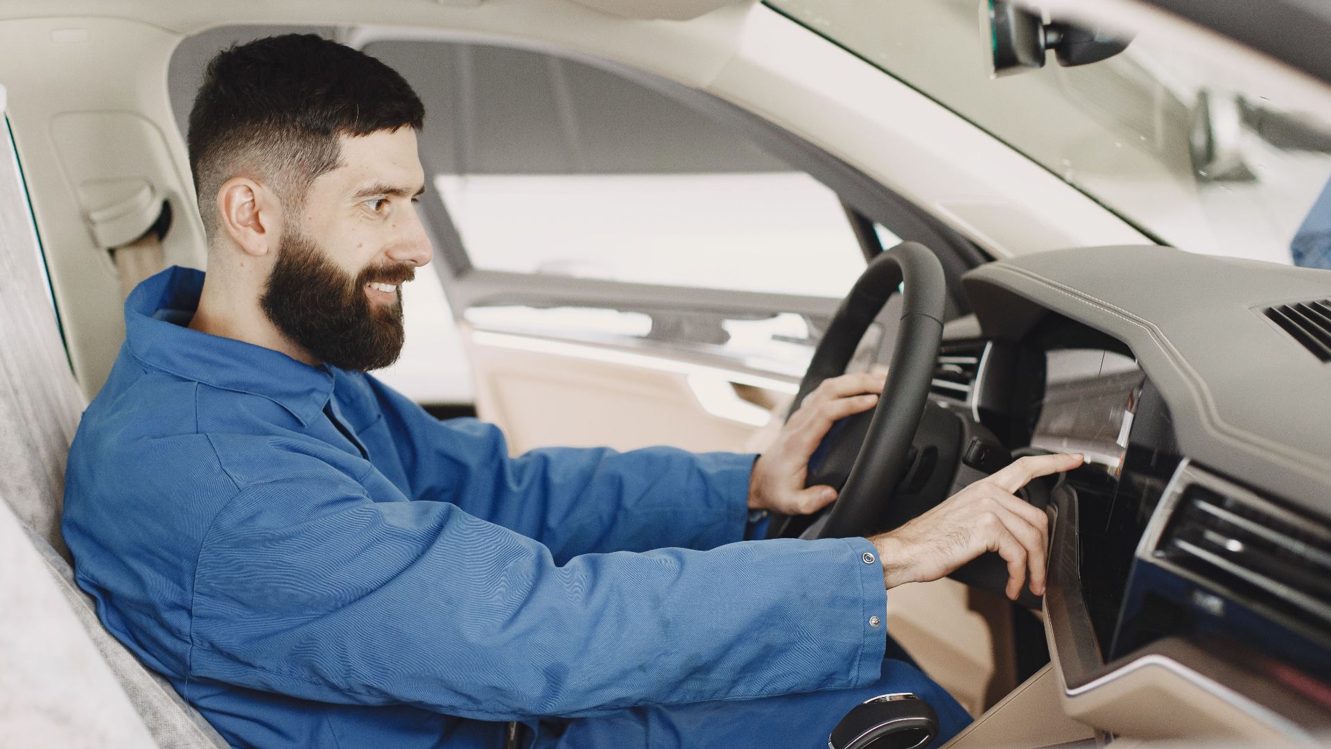Mechanic in blue coveralls interacts with car dashboard, smiling and focused.