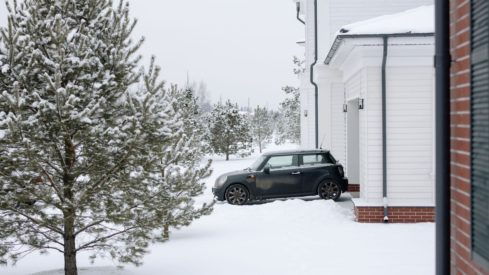A Mini Cooper parked in a snowy driveway beside a white house during winter.