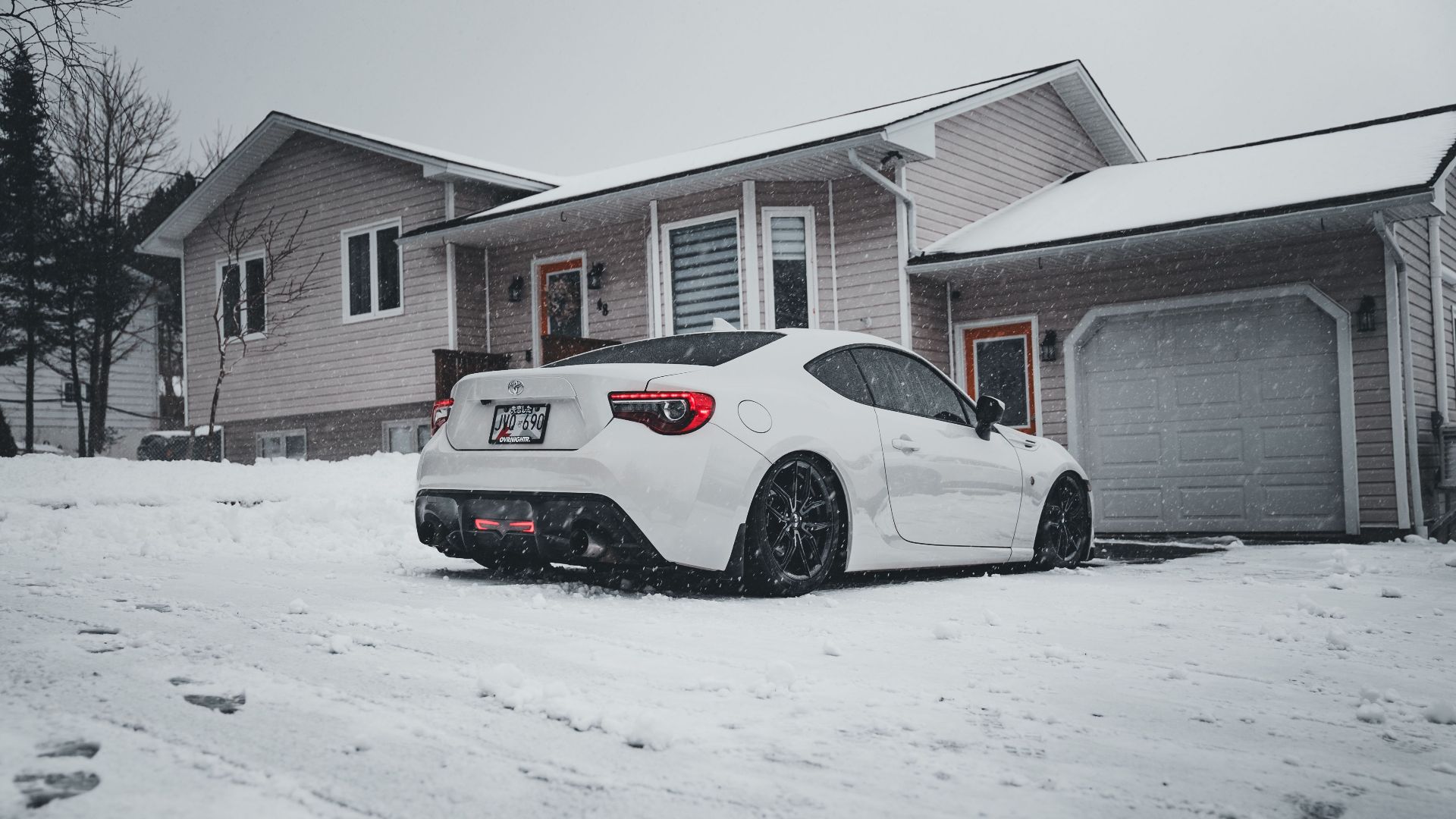 A white sports car parked in a snowy suburban driveway during winter snowfall.