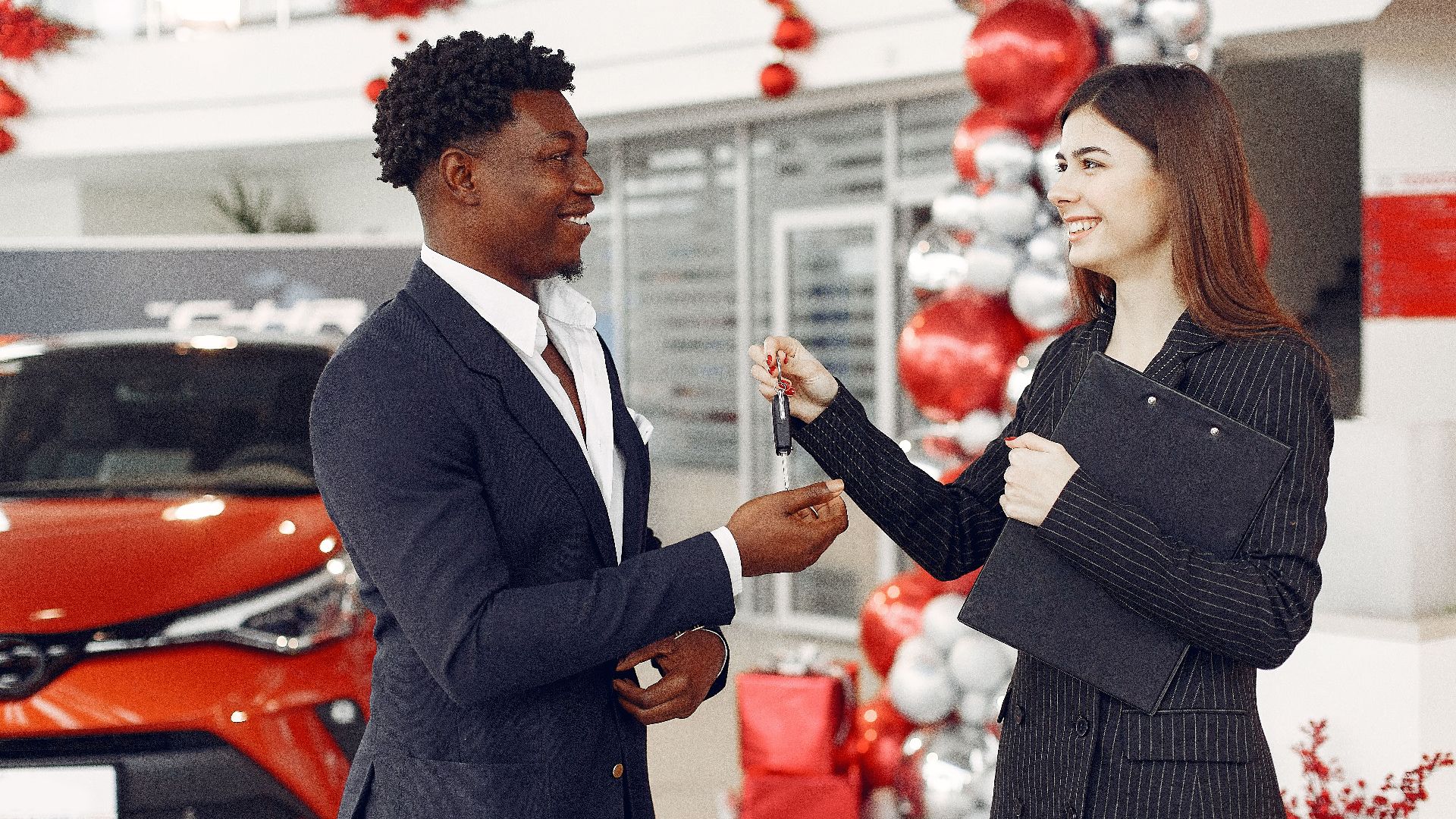 Side view of happy positive female dealer in formal classy wear passing car keys to  excited African American customer in trendy suit in car showroom
