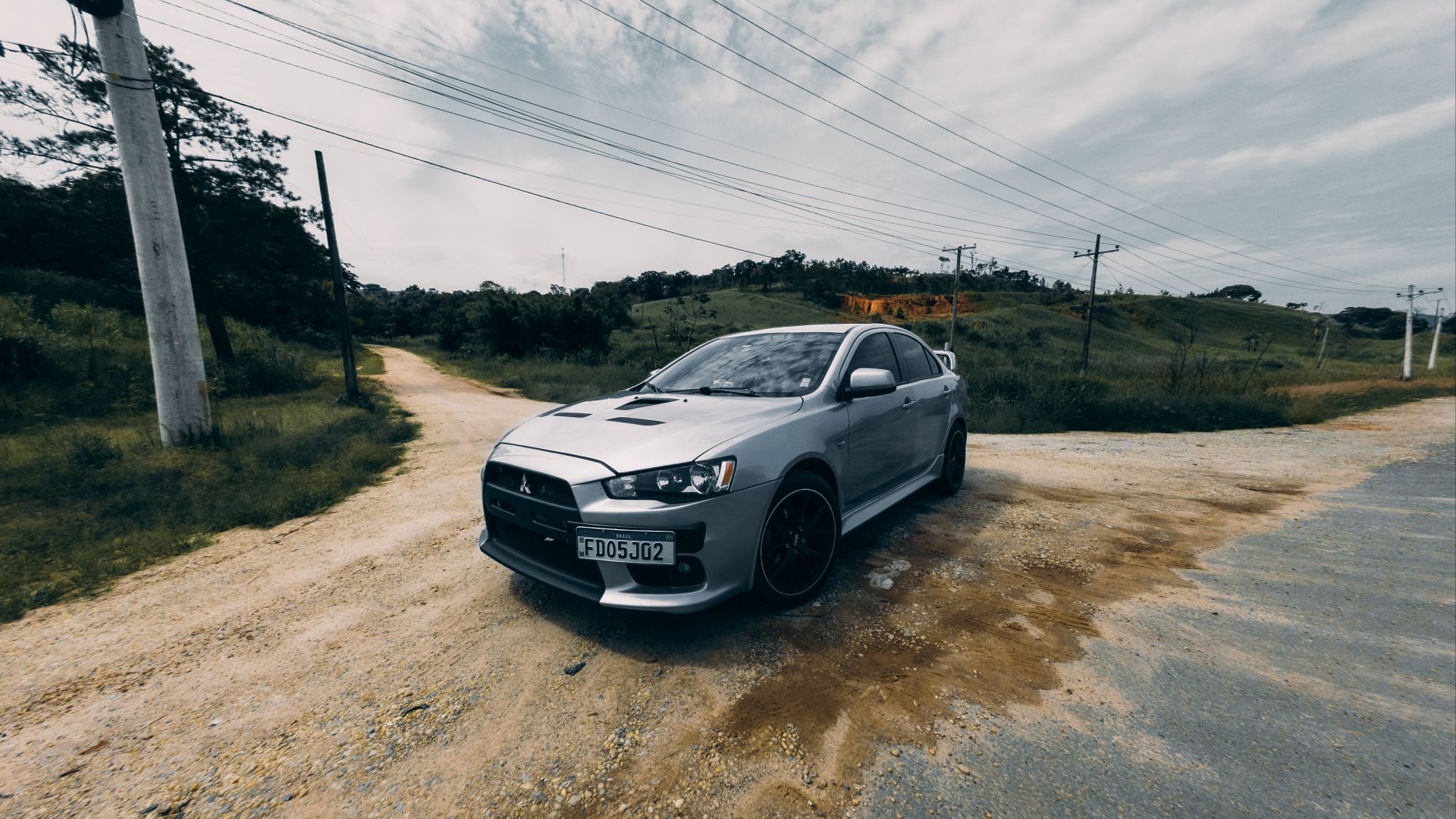 A gray car parked on a rural dirt road intersection with distant hills and power lines.