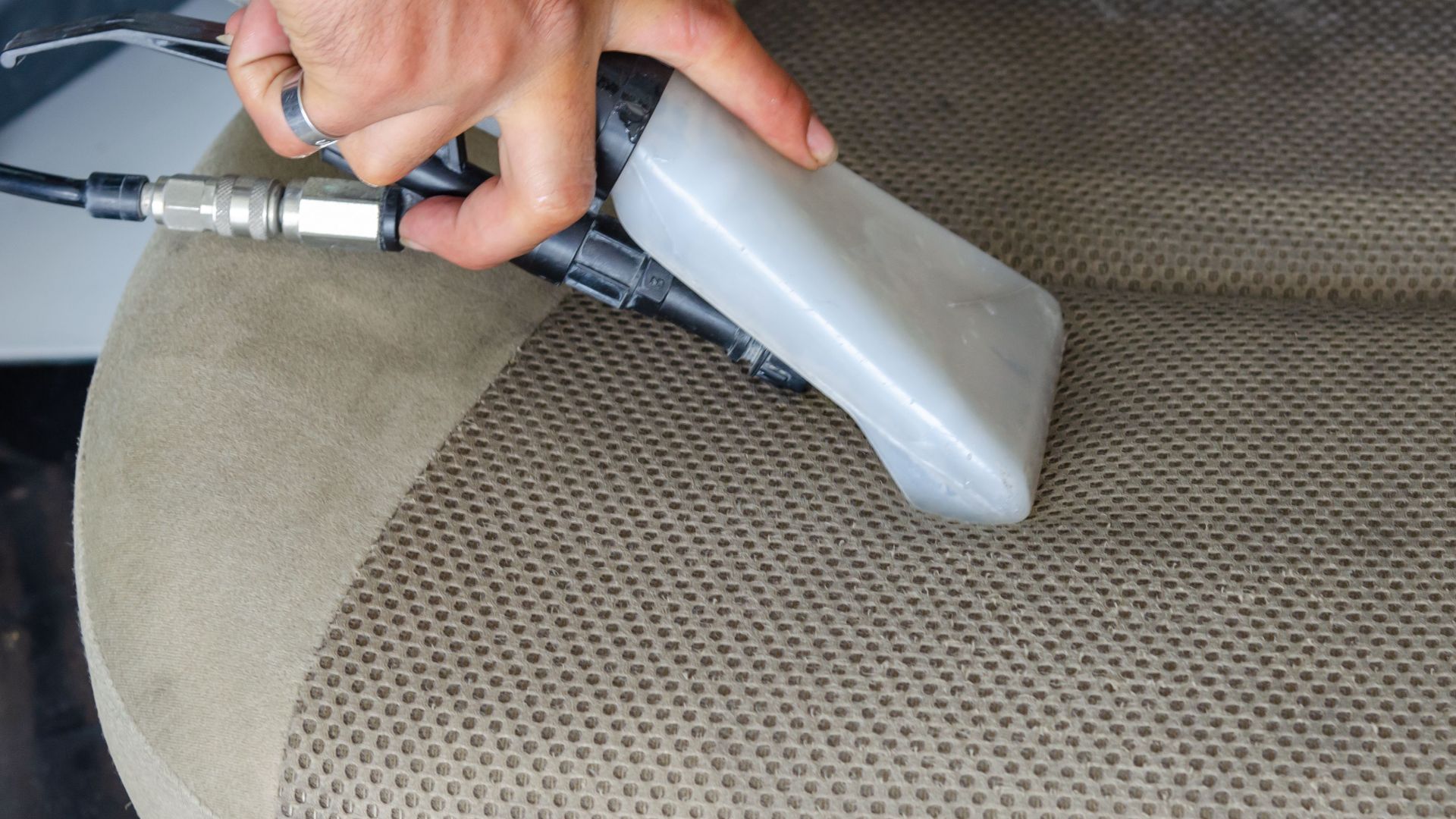 Hands using a vacuum nozzle to deeply clean a fabric car seat, removing dirt and stains.