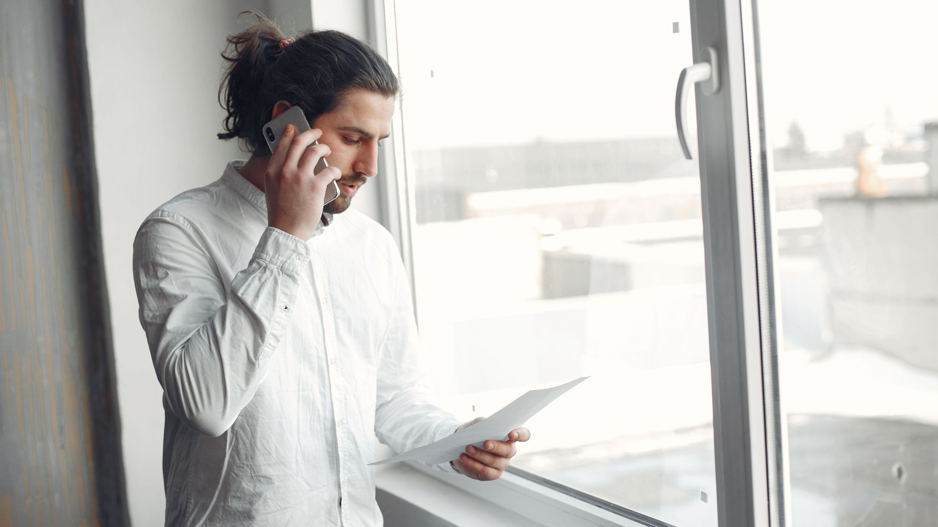 Young man in white shirt, on phone call holding a document, standing by a large window.