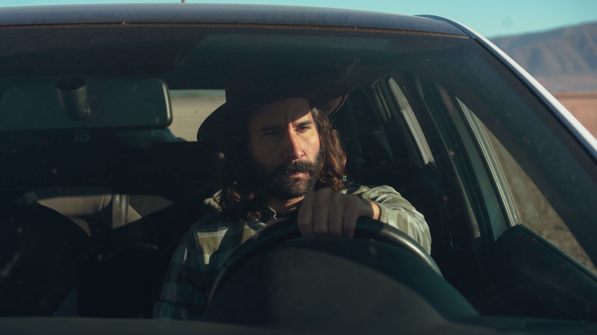 A bearded man in a hat driving a car through a sunny desert landscape, conveying a sense of adventure.