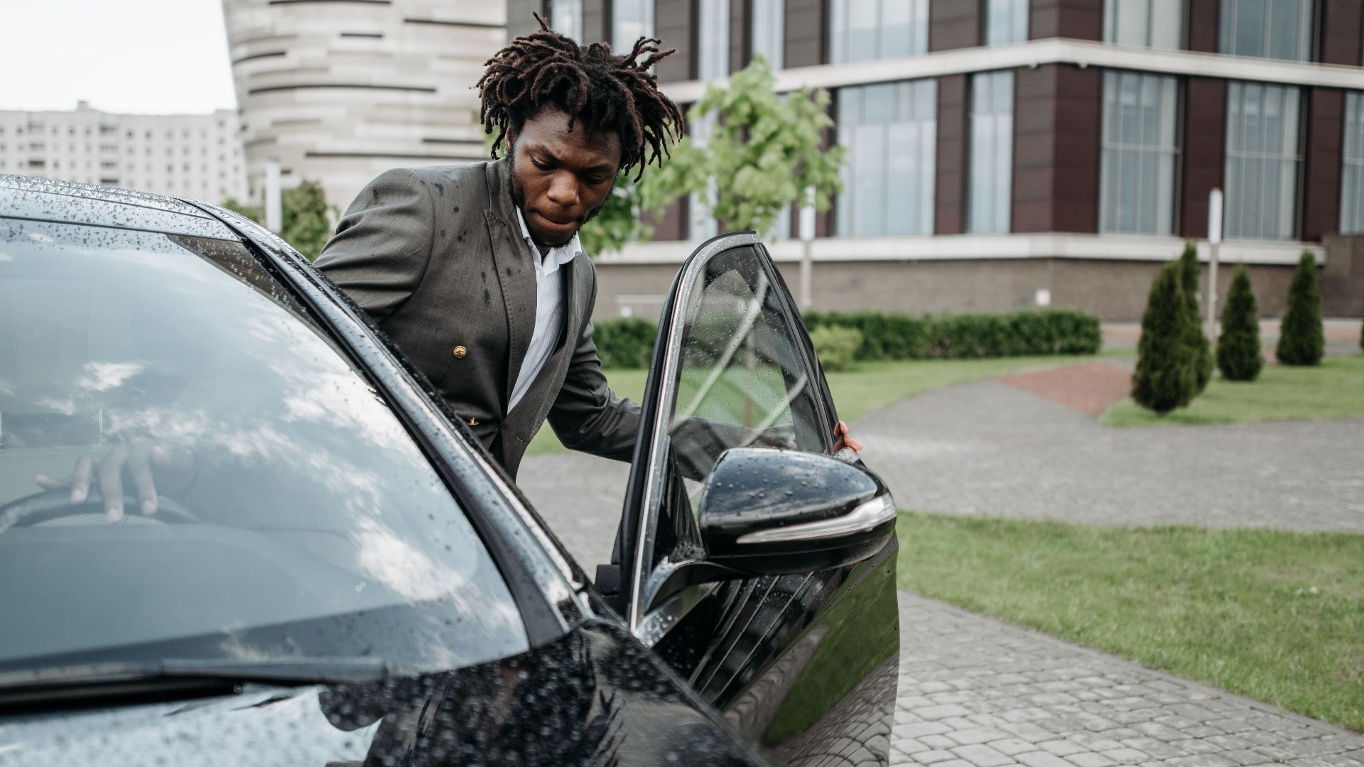 An African American man in a gray suit exits a black car in front of a modern office building on a rainy day.