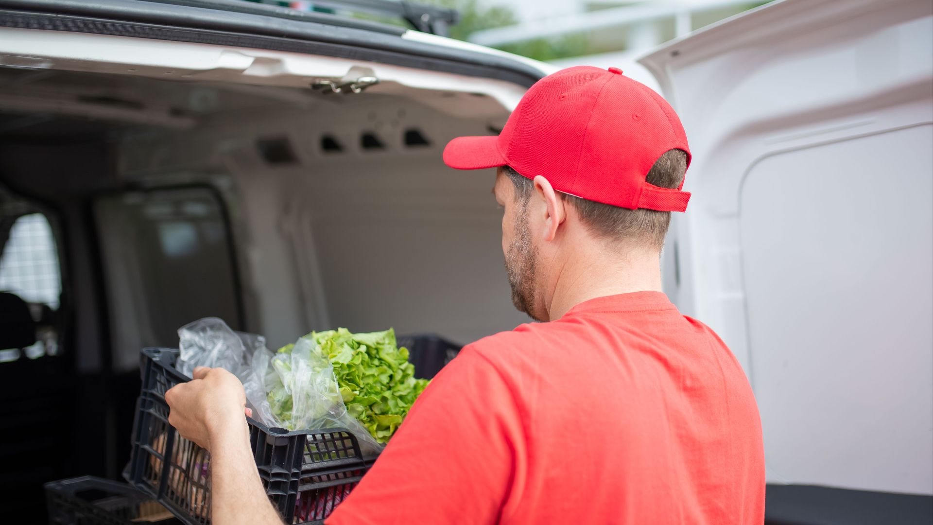 A delivery man in red loading groceries into a van, focusing on fresh produce.