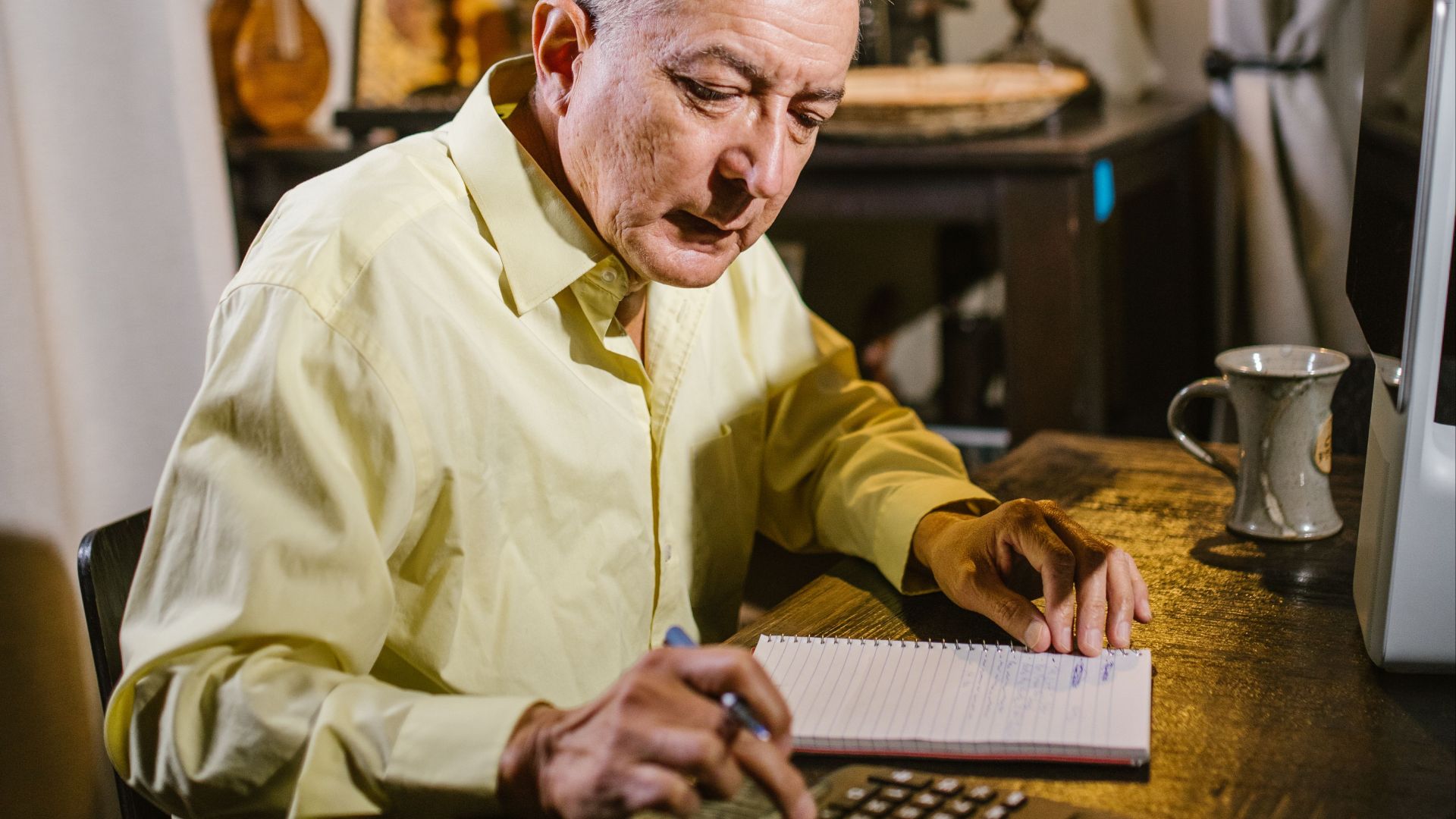 Elderly man working on calculations and taking notes in home study with warm lighting.