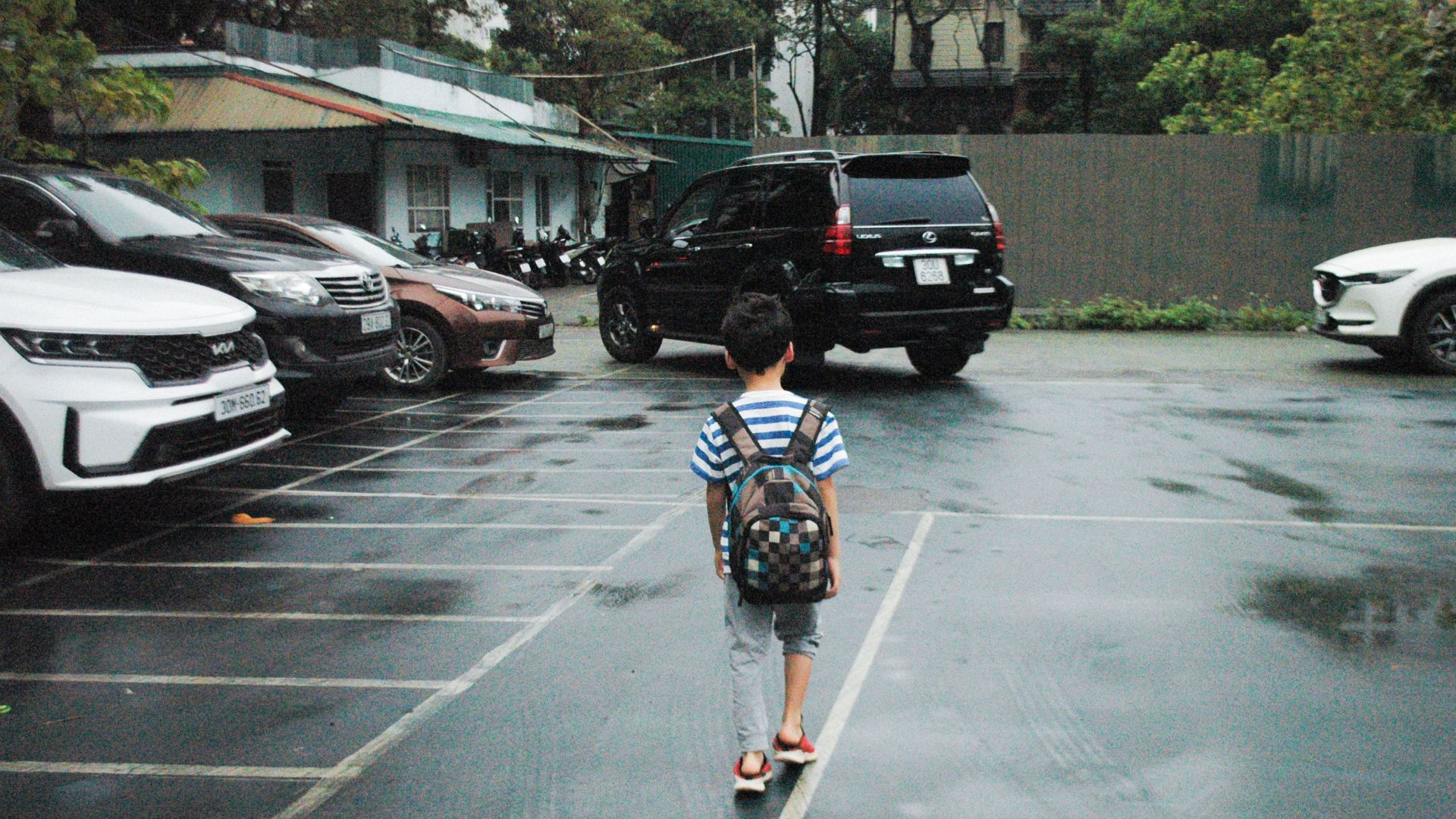A young boy with a backpack walks through a parking lot, surrounded by cars on a rainy day.