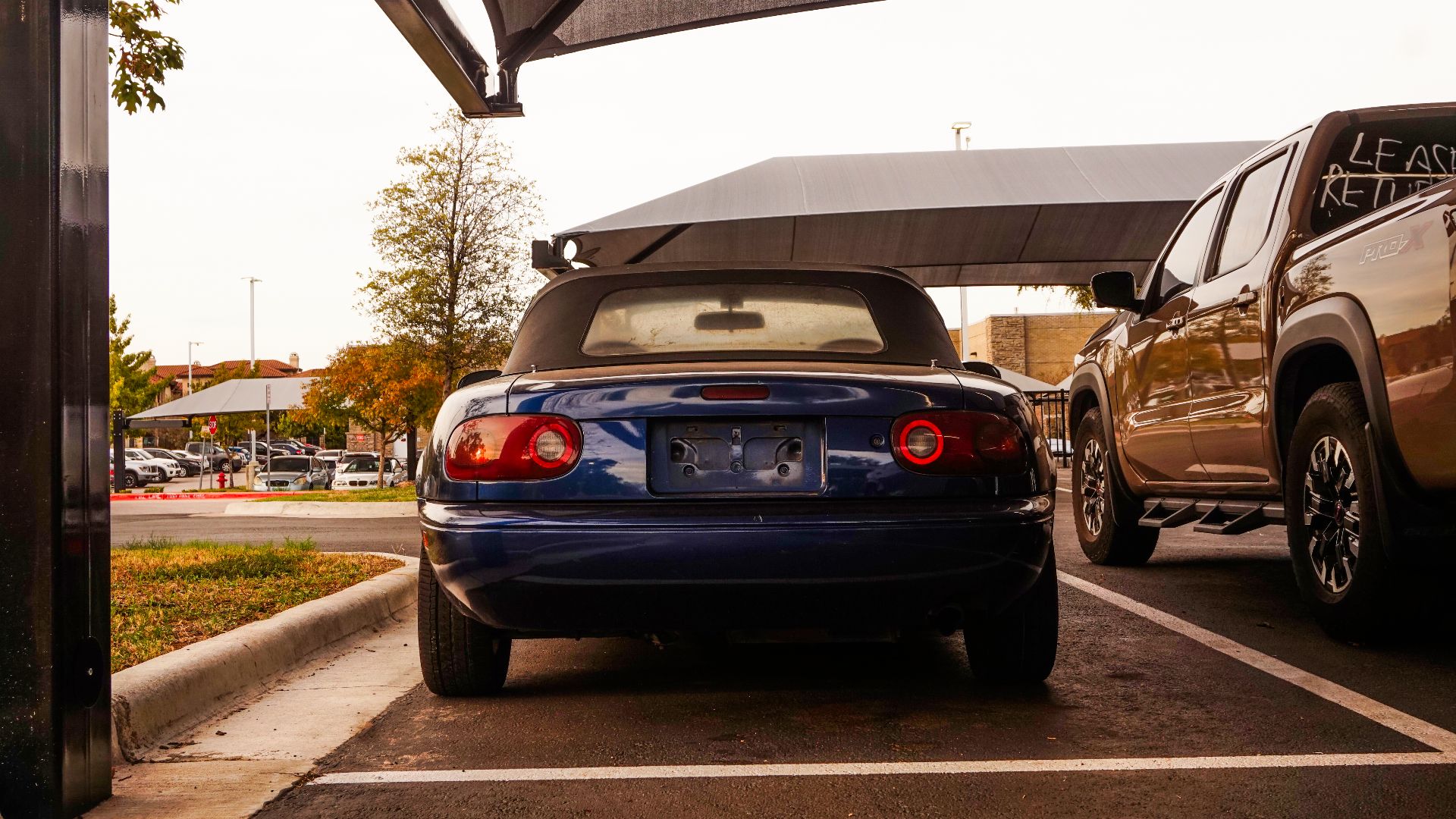 Rear view of a vintage blue convertible car parked beside a pickup in a city parking lot.
