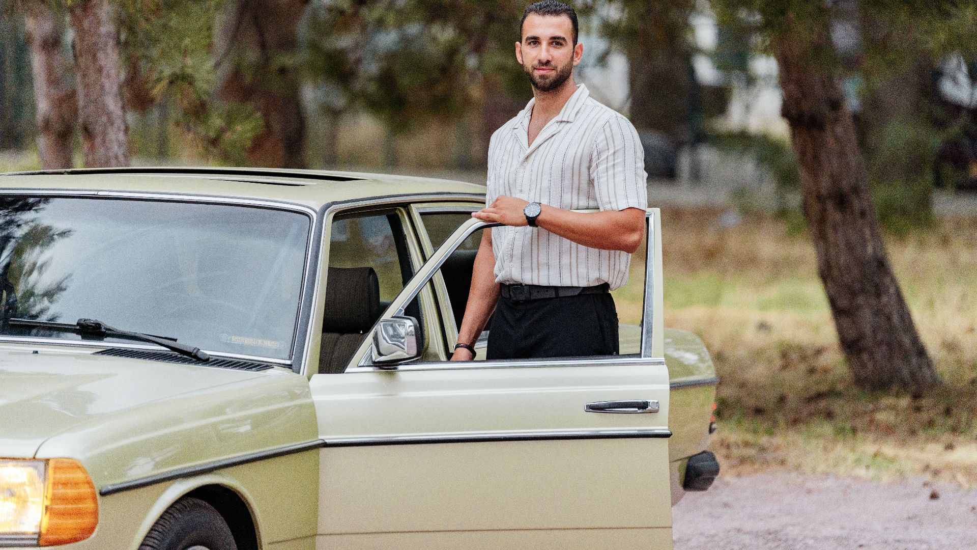 A man stands by an open door of a classic green car, outdoors among tall trees.