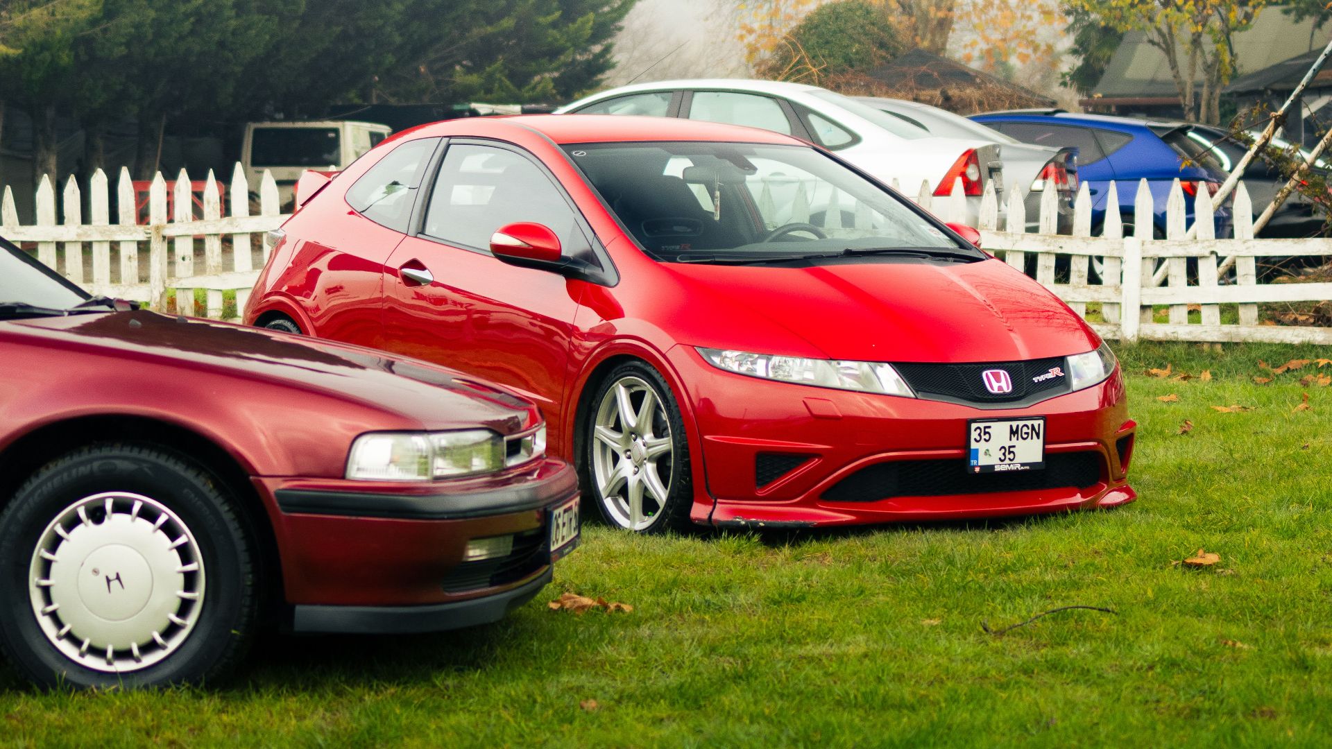 Two red cars parked on a grassy yard with white wooden fences in a suburban setting.