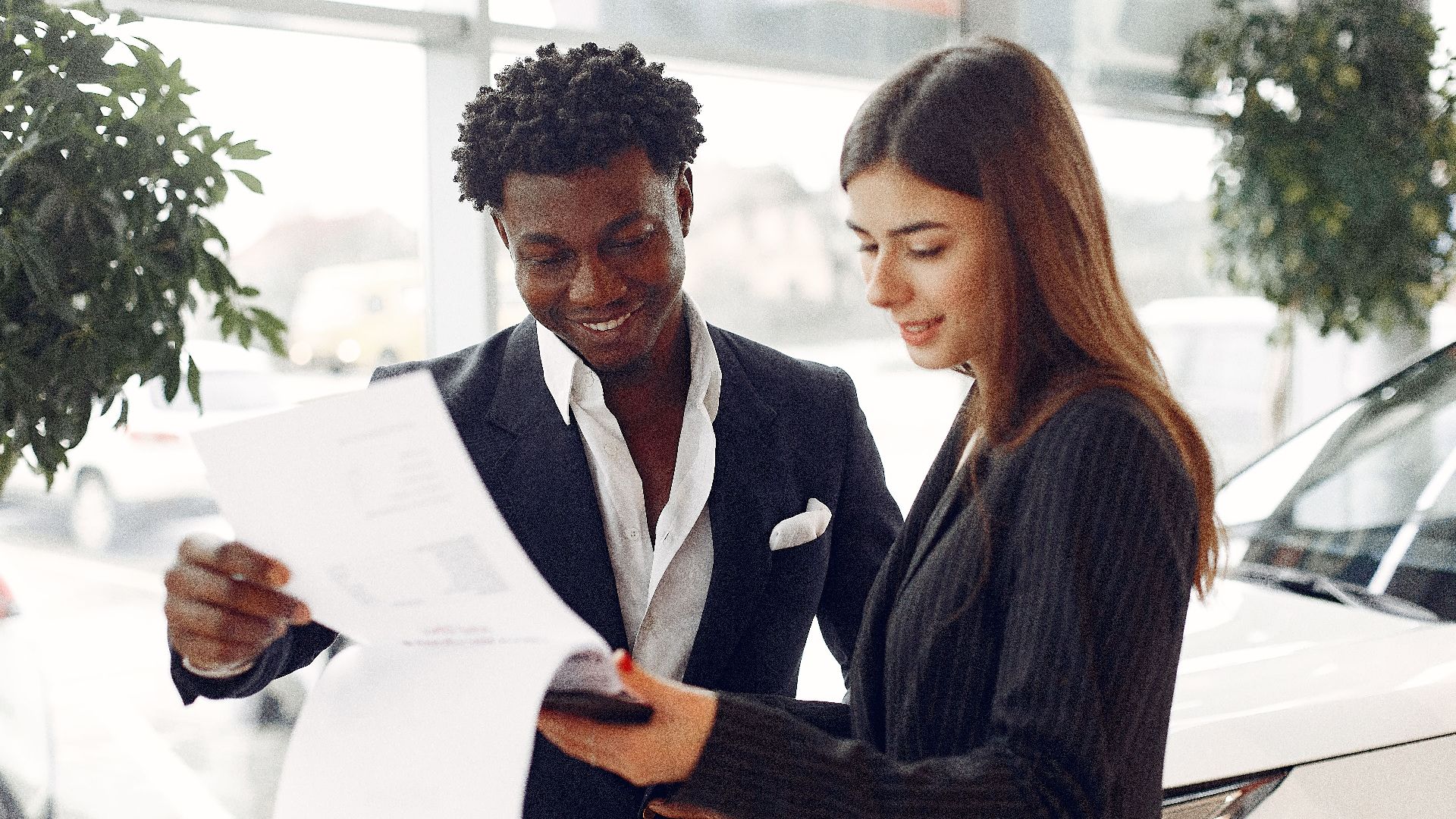 Cheerful smiling multiethnic businessman in classy suit and professional elegant female consultant standing close and reading contract details in car showroom