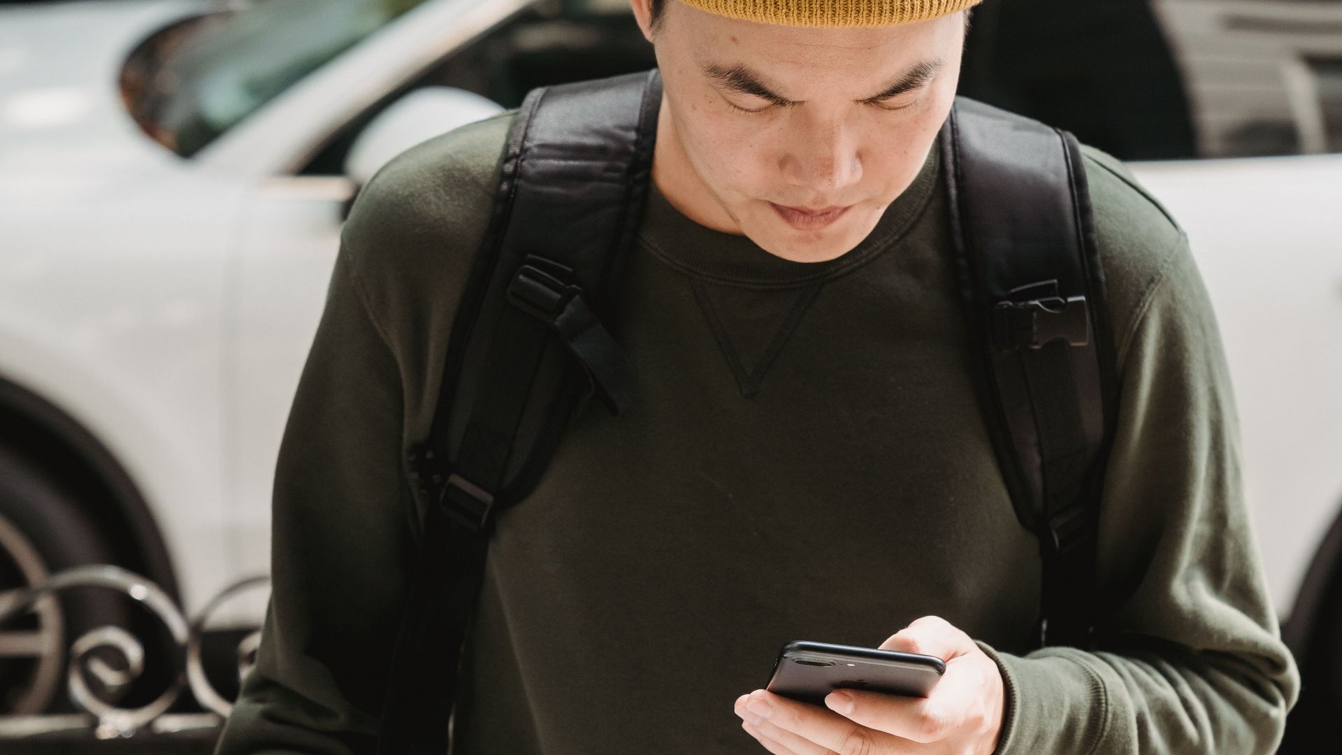 Asian deliveryman in beanie checks phone while carrying package outdoors.