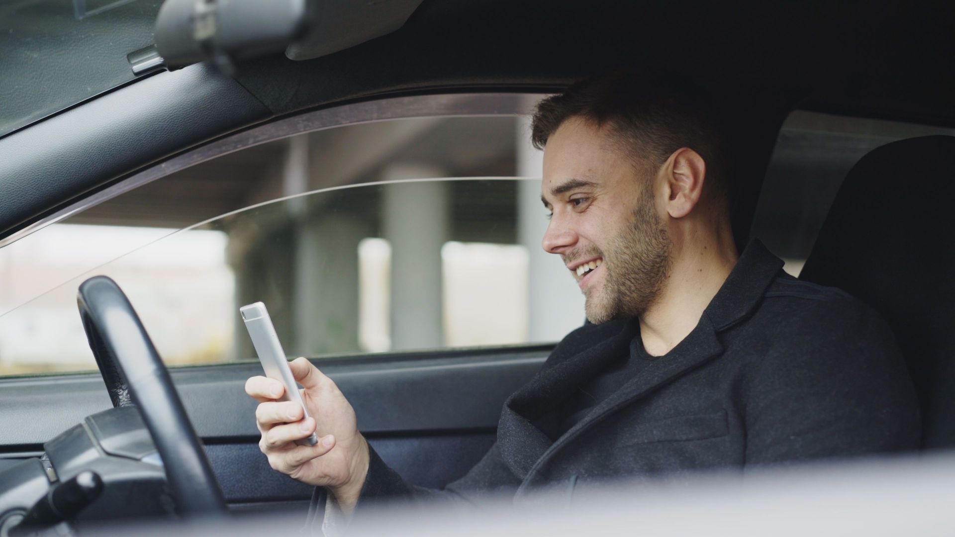 Adult man smiling while using a smartphone inside a car. Relaxed and happy expression.