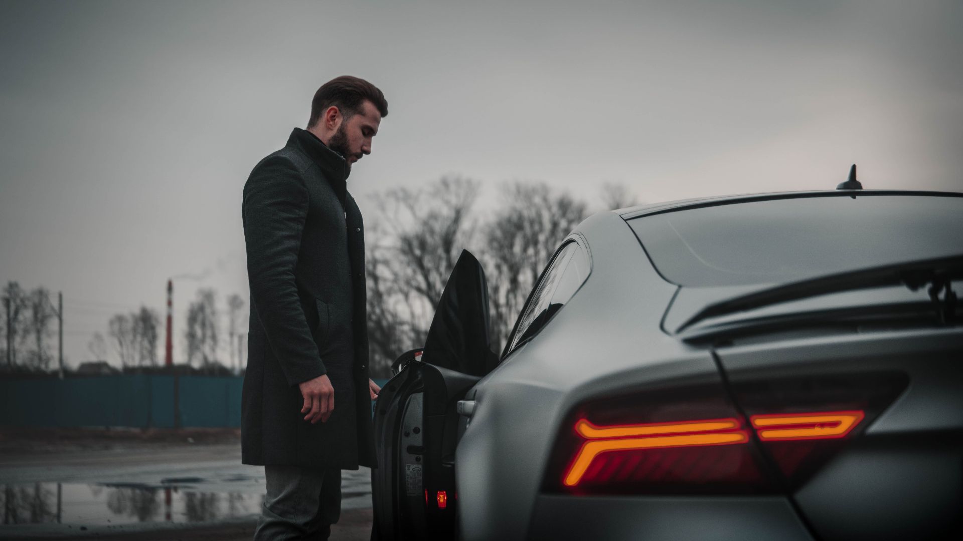 A man in a black coat stands beside a luxury car on a cloudy day in Moscow, Russia.