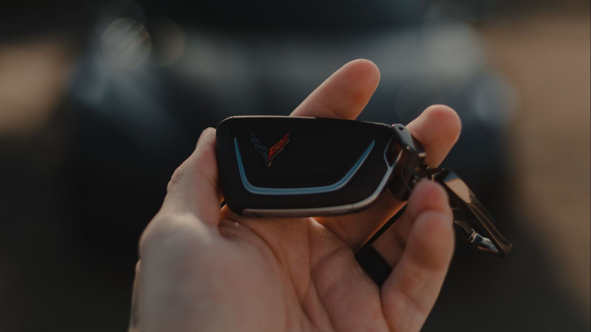 A hand holding a car key fob with a blurred car in the background, outdoors.
