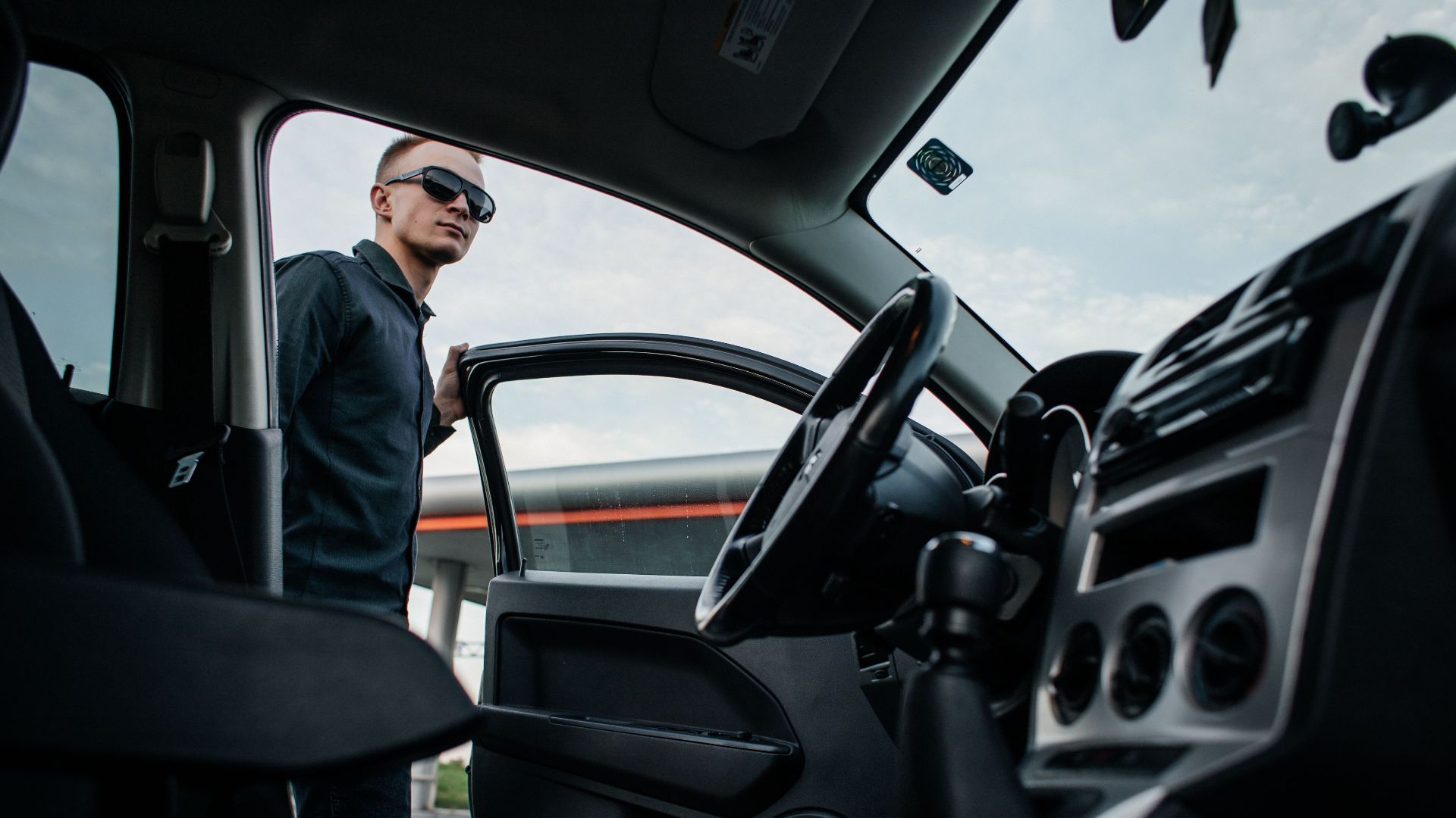 A man in sunglasses stands by an open car door, viewed from the interior.