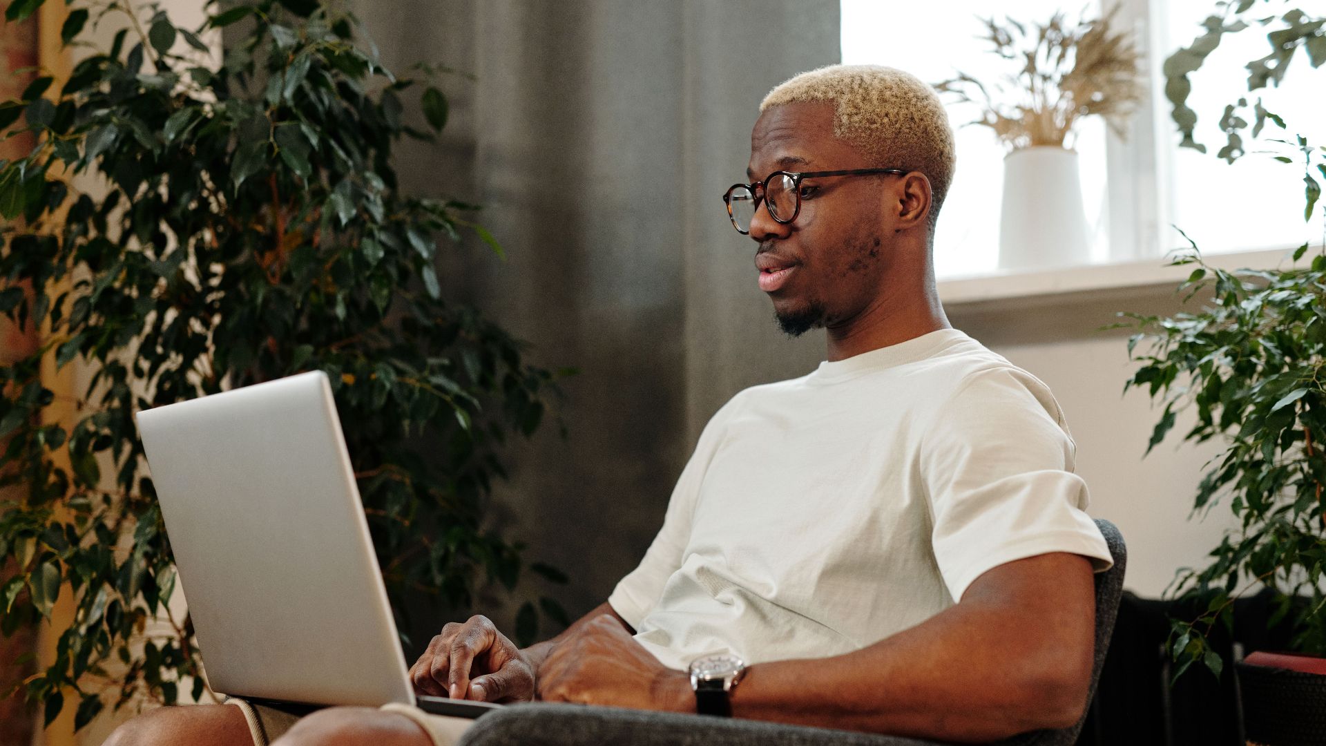 Focused young man with glasses working on a laptop in a cozy indoor space surrounded by plants.
