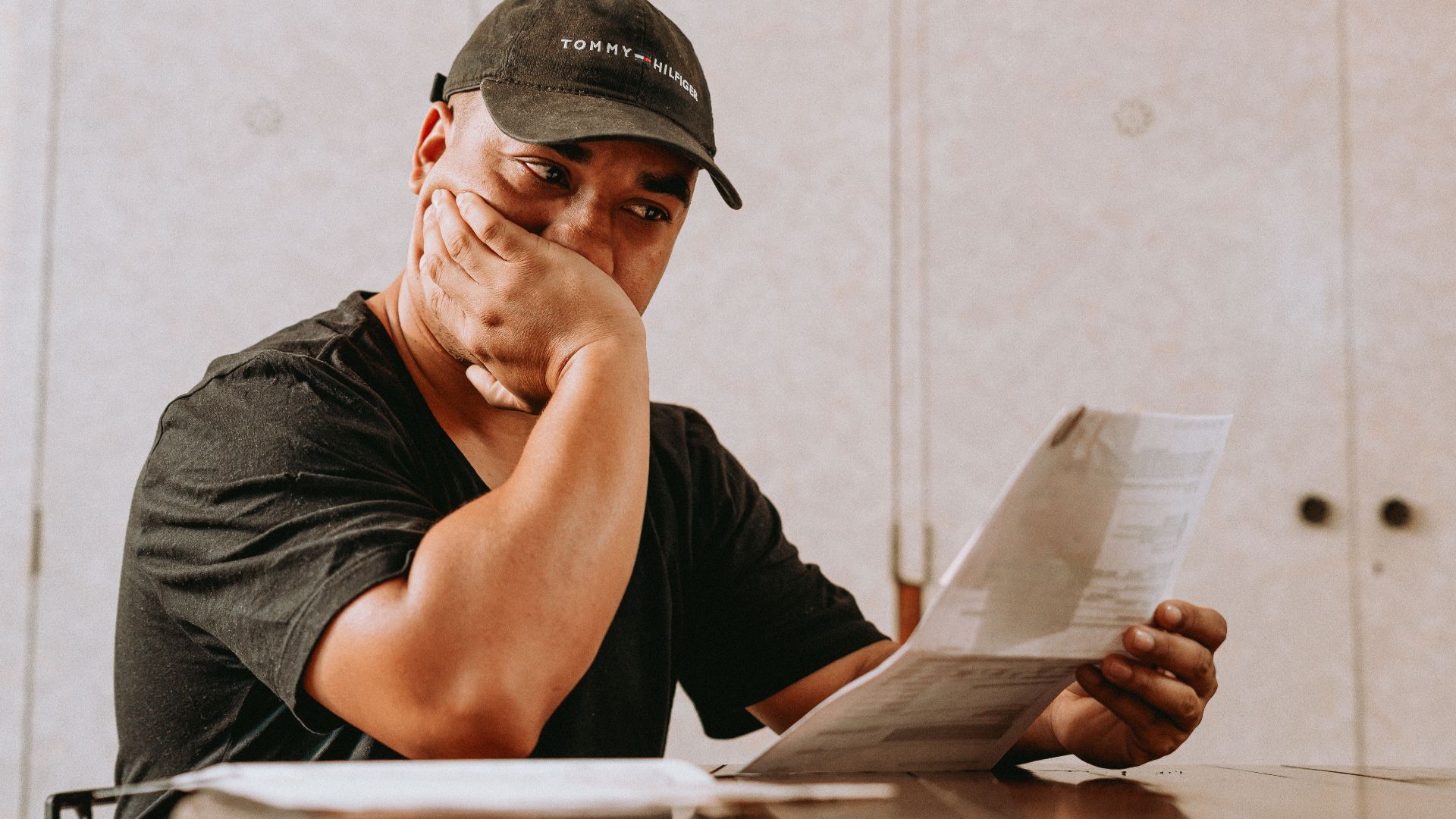 A man sits indoors, deeply focused on reviewing paperwork, expressing concern and contemplation.