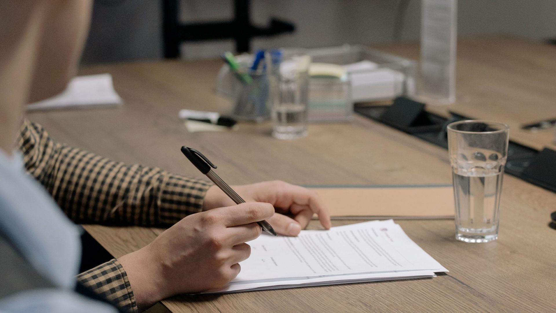 Person in business attire signing a document at a wooden table in an office setting.