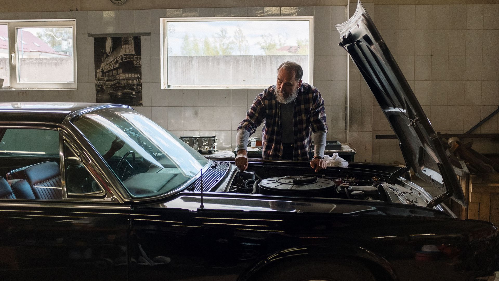 A bearded mechanic examines a classic car engine in a well-lit auto repair shop.