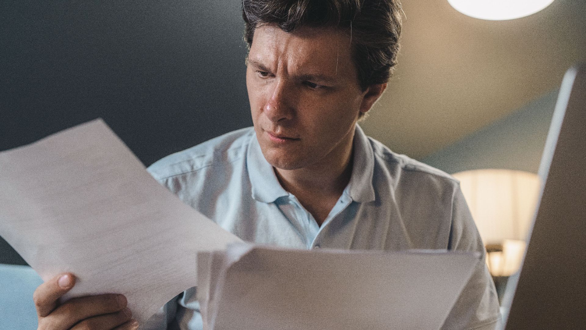 A man intently examines papers, seated indoors under warm lighting, focusing on his work.