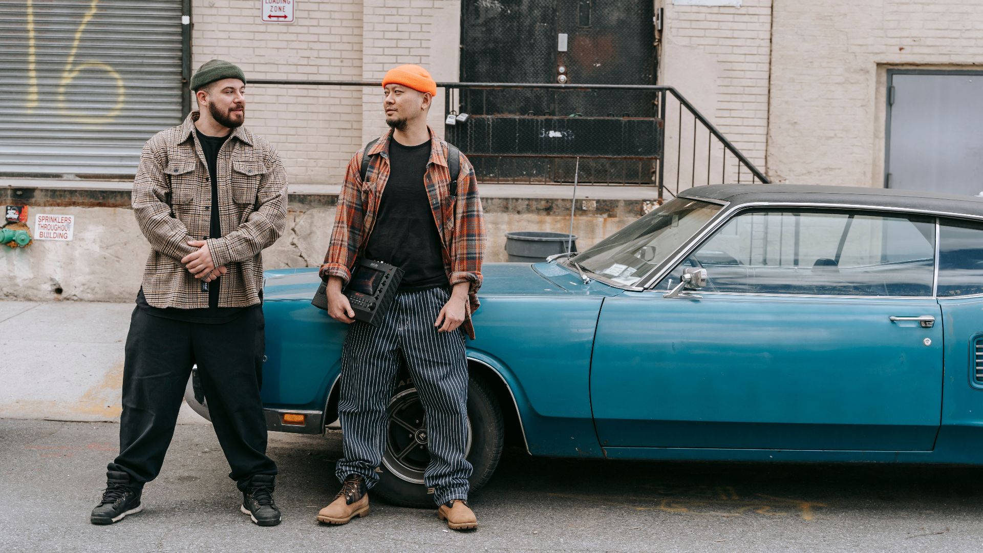Two men in casual urban outfits stand beside a vintage blue car on a city street.
