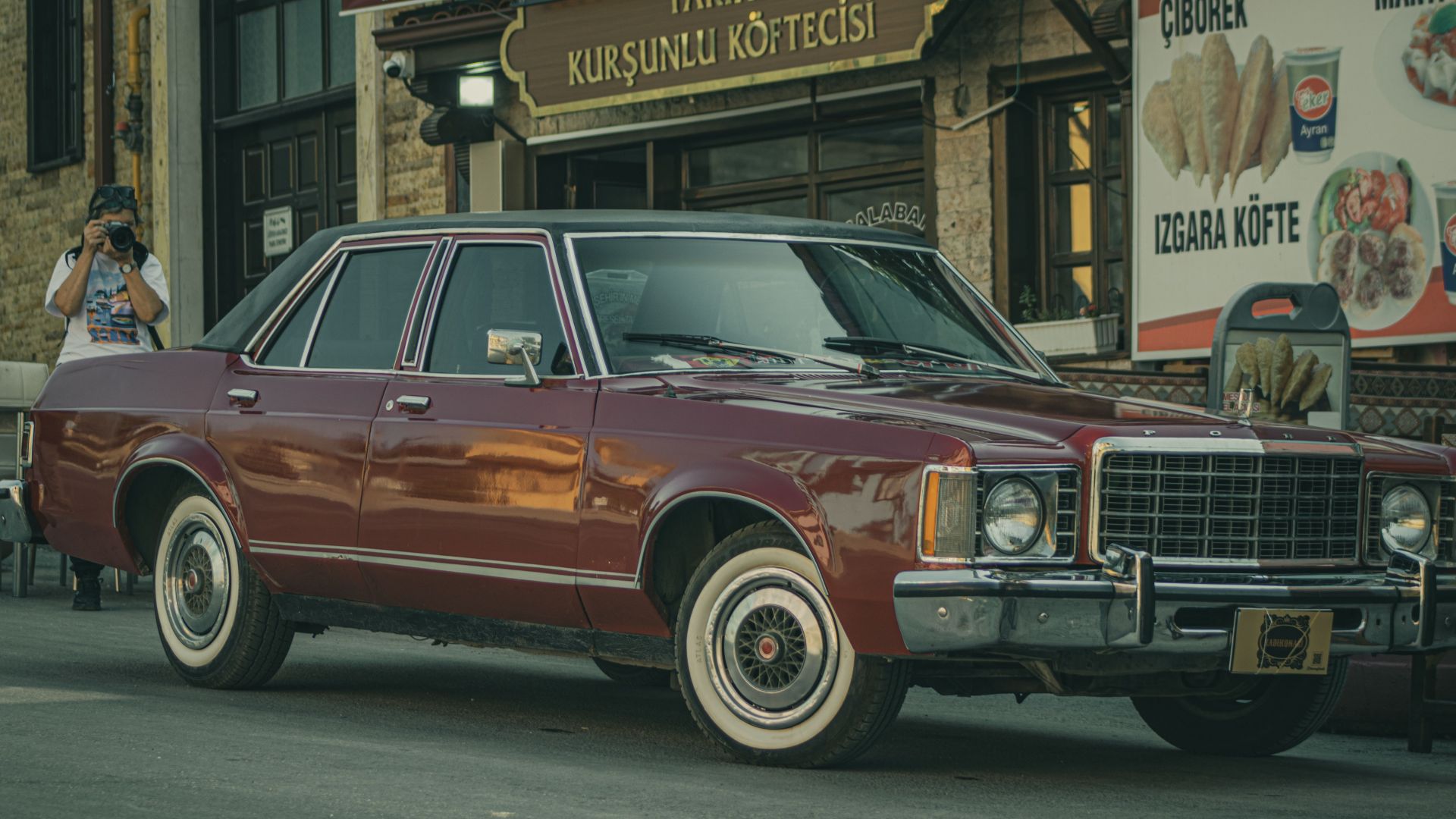Classic vintage car parked outside a Turkish restaurant with food signs and photographer in view.