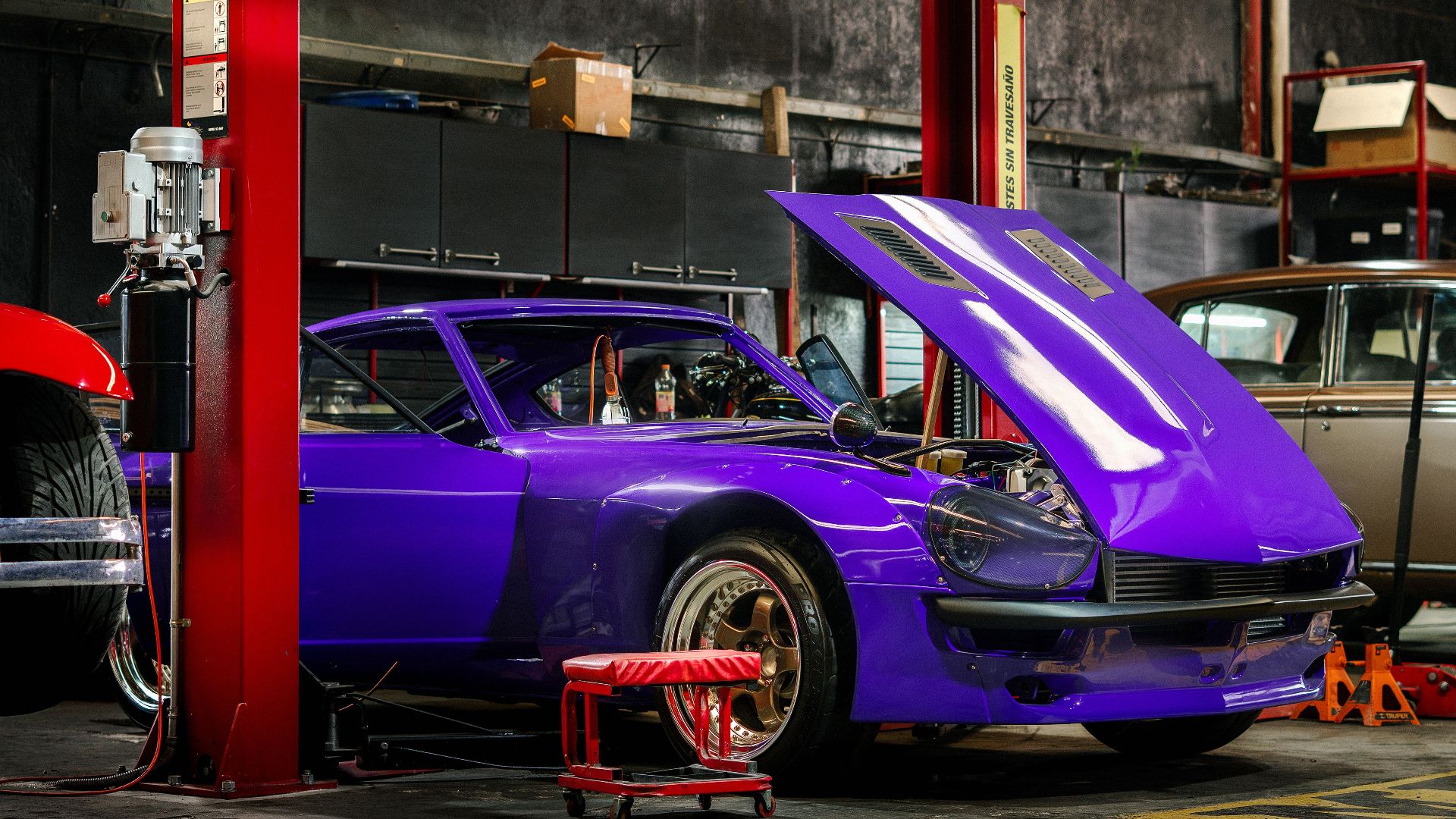 A classic car with a vibrant purple color in a mechanical workshop with its hood open.
