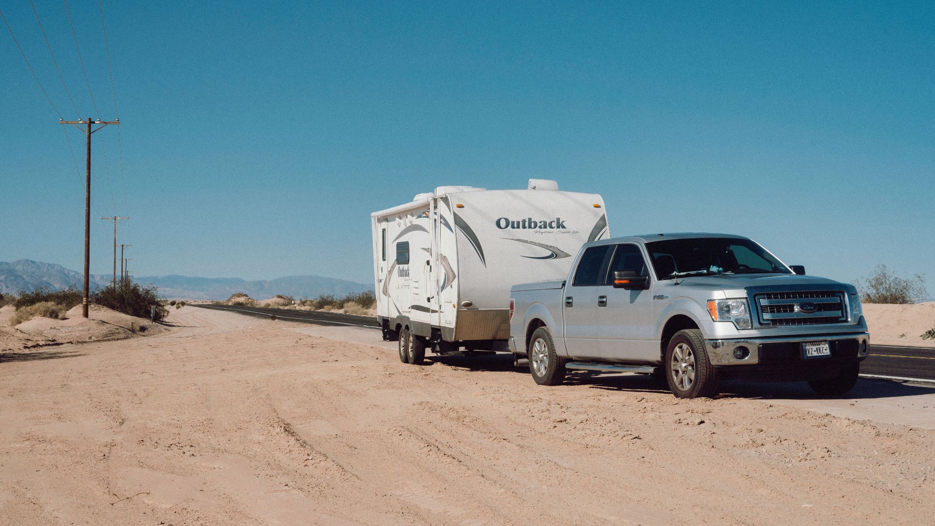 Silver truck towing an Outback trailer on a desert road under clear blue skies.