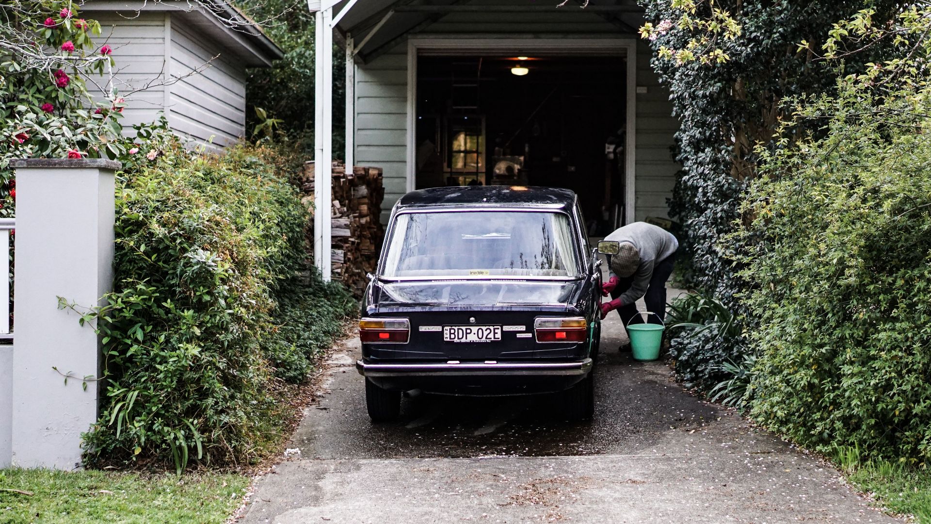 A classic automobile parked in a lush suburban driveway with a person cleaning it in Leura, NSW.