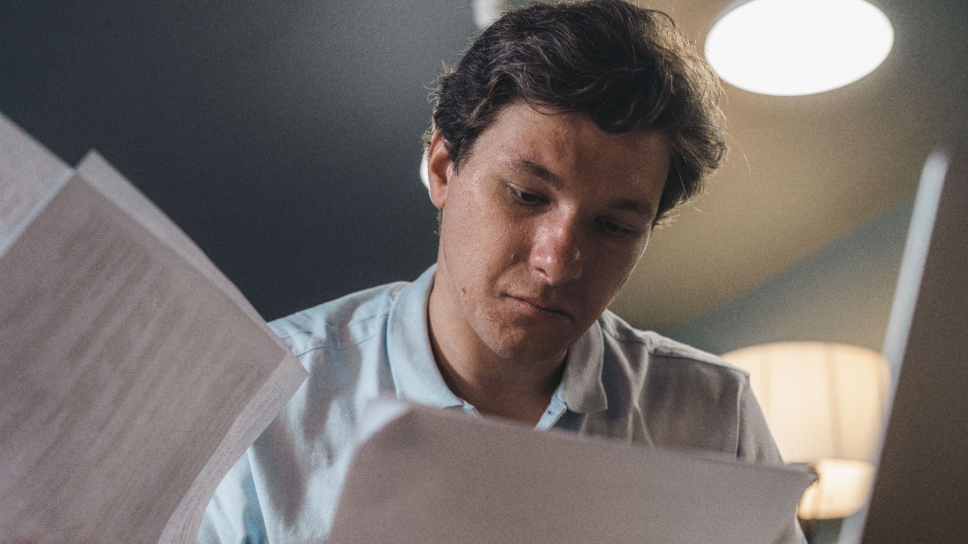 Serious man wearing a light blue shirt intently reading printed papers indoors under warm lighting.