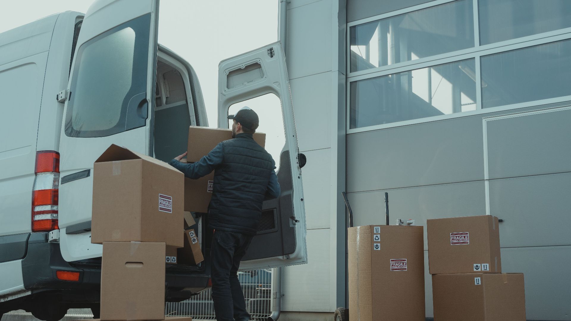 A delivery man unloading cardboard boxes from a van at a warehouse during the day.
