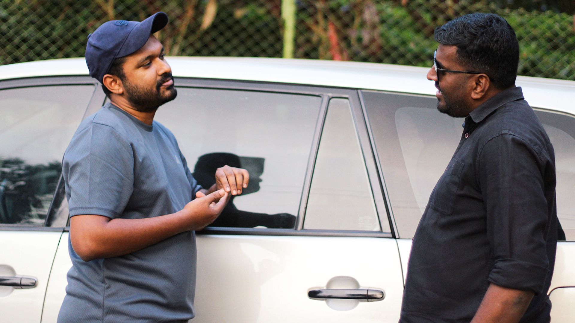 Two men having a casual discussion beside a car in an outdoor setting.