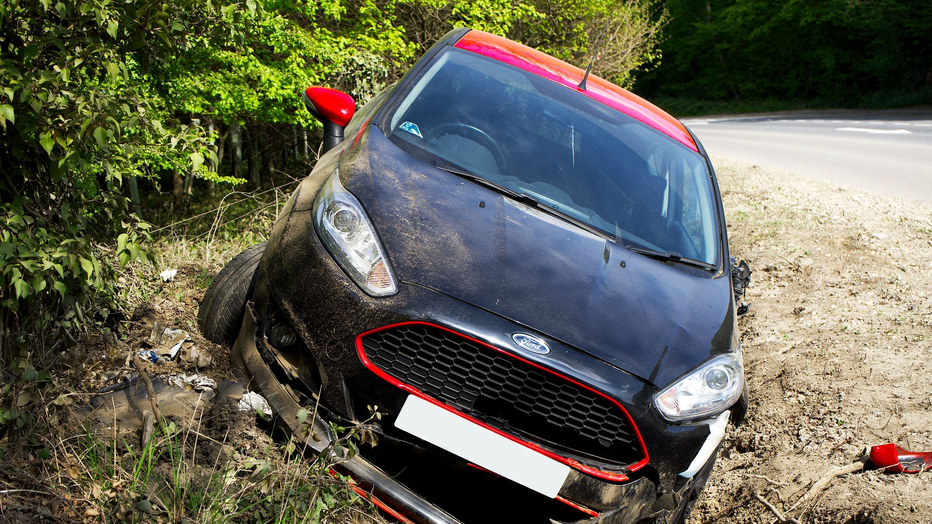 A black car, seemingly a Ford, crashed and damaged in a roadside ditch with surrounding greenery.