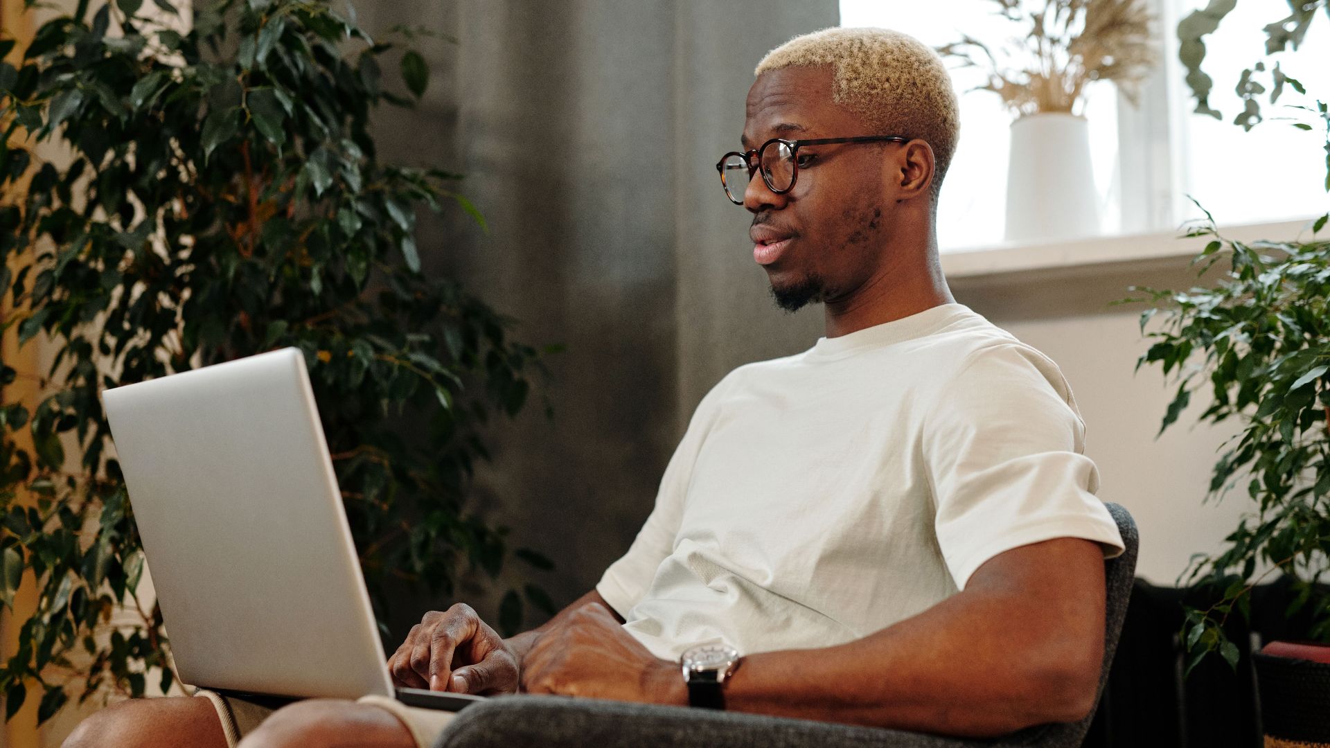 Focused young man with glasses working on a laptop in a cozy indoor space surrounded by plants.