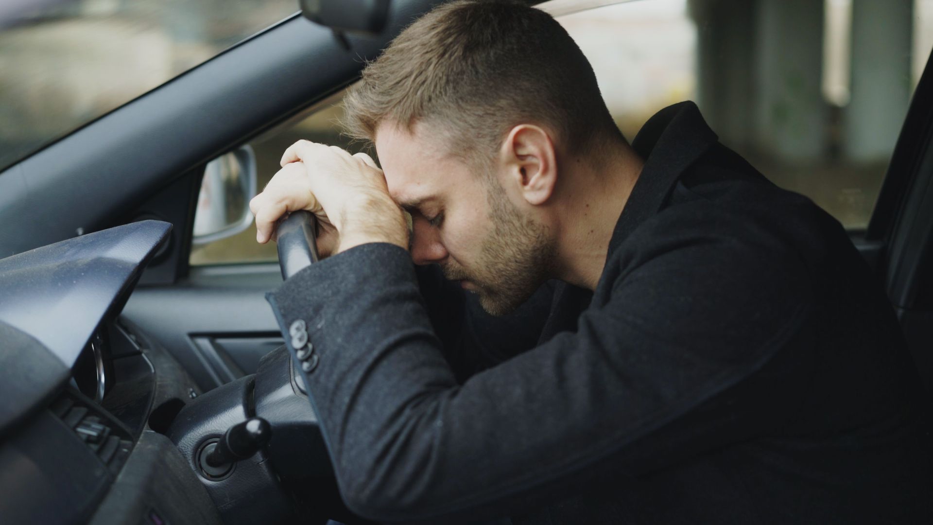 A man showing stress while sitting in a car, head on steering wheel.