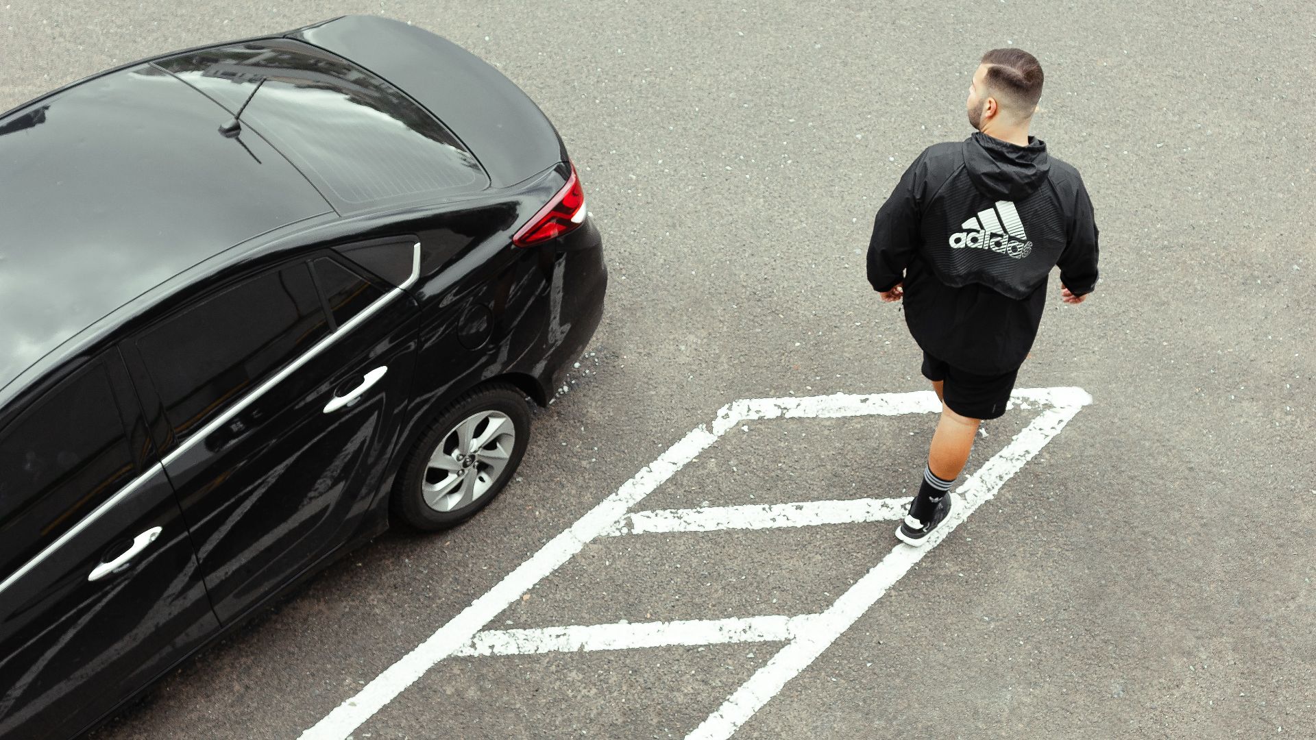 Aerial image of a man in sportswear walking near a parked black car in an empty asphalt lot.